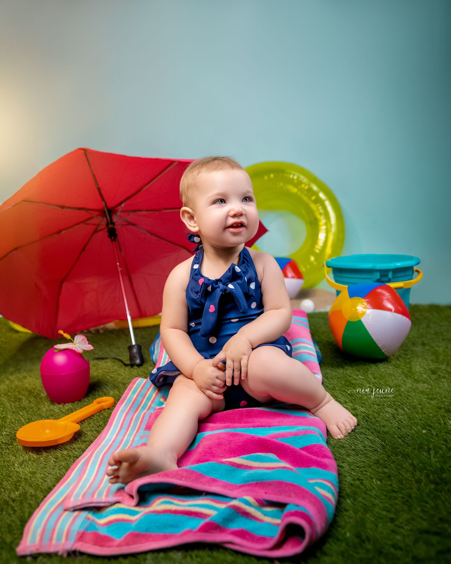 Baby in blue outfit poses with colorful beach toys and umbrellas on artificial grass.