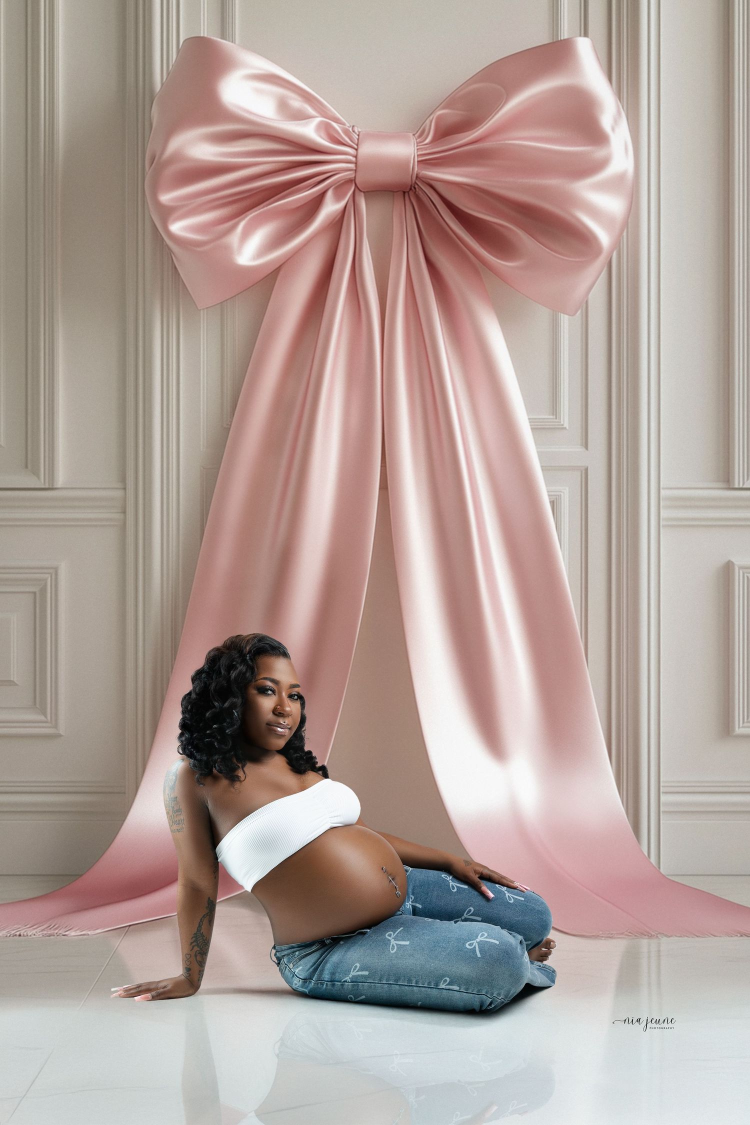A maternity photo featuring a large pink satin bow backdrop and white paneled walls.