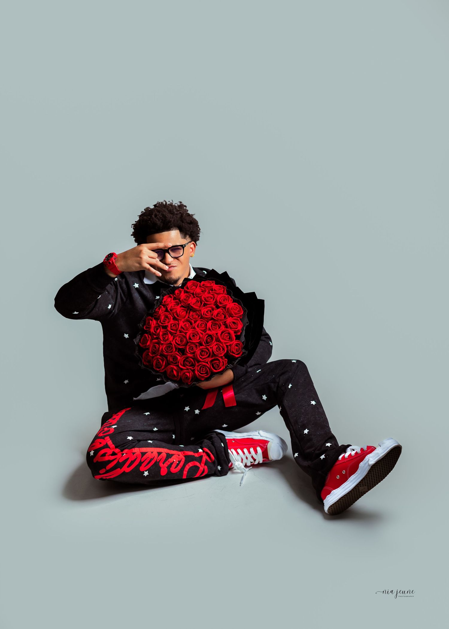 Young man sitting on floor holding red roses bouquet, wearing black outfit with red sneakers against gray background.