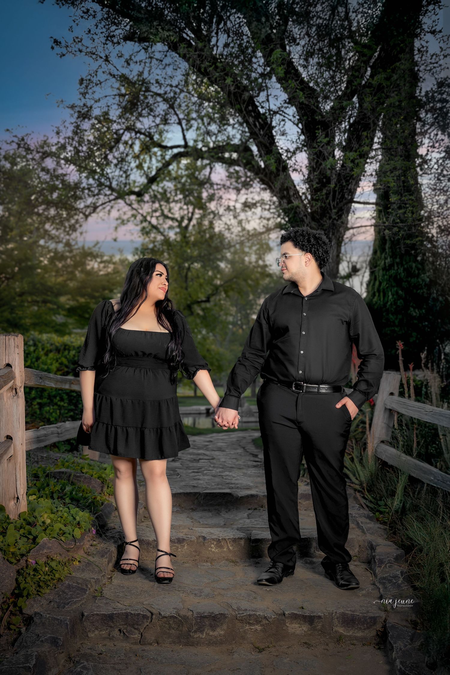 A couple in black attire holds hands while standing on a rustic bridge path during sunset.