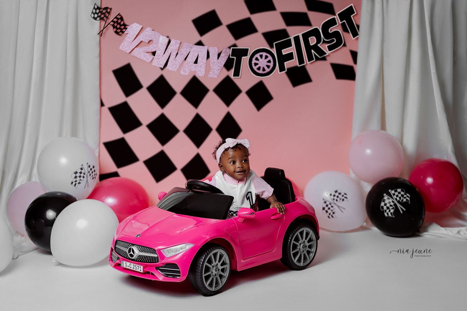 Pink Mercedes ride-on toy car against pink backdrop with black and white checkered banner and balloons.