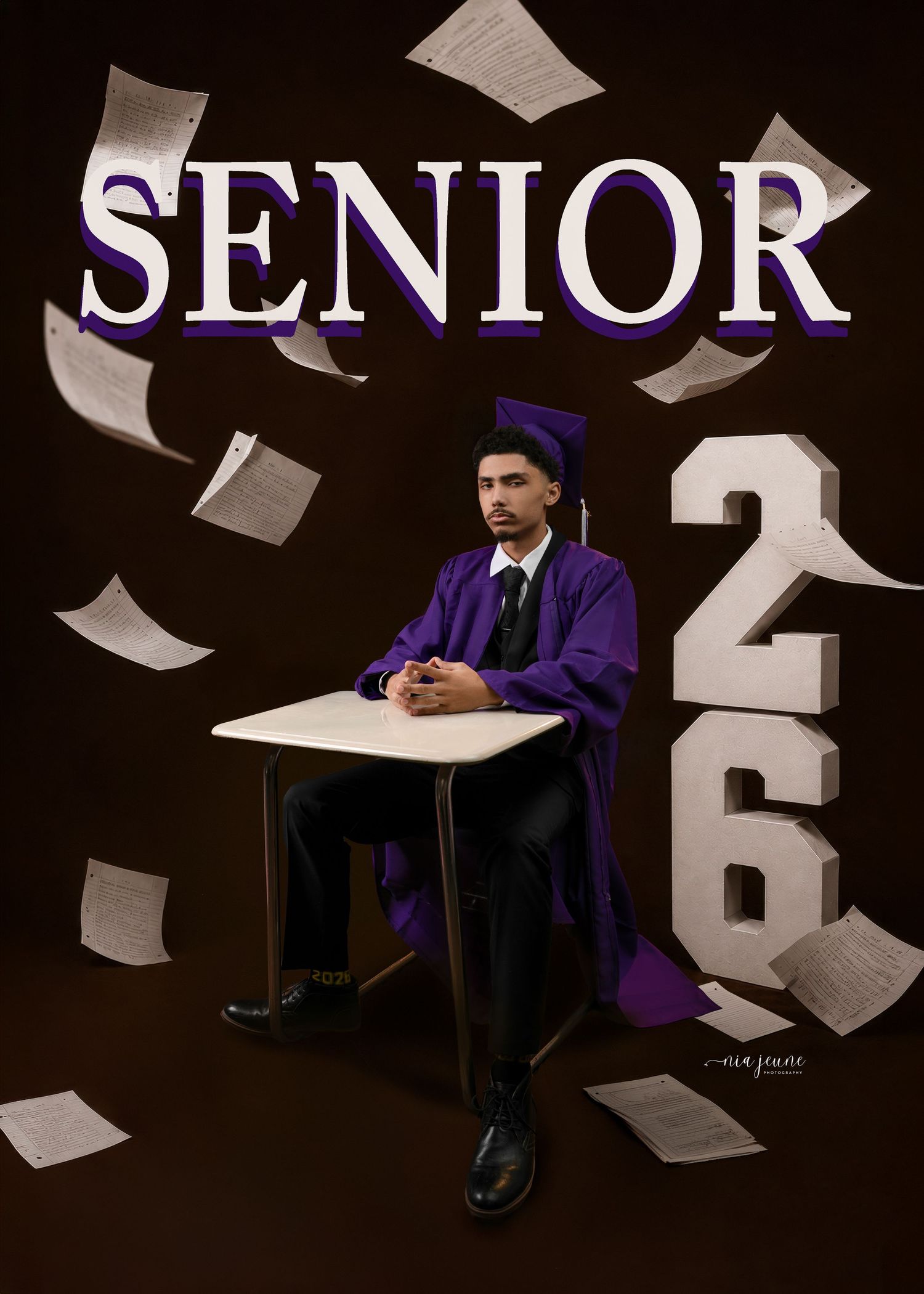 Senior 2026 graduate in purple cap and gown sitting at desk with flying papers in dramatic dark studio portrait.