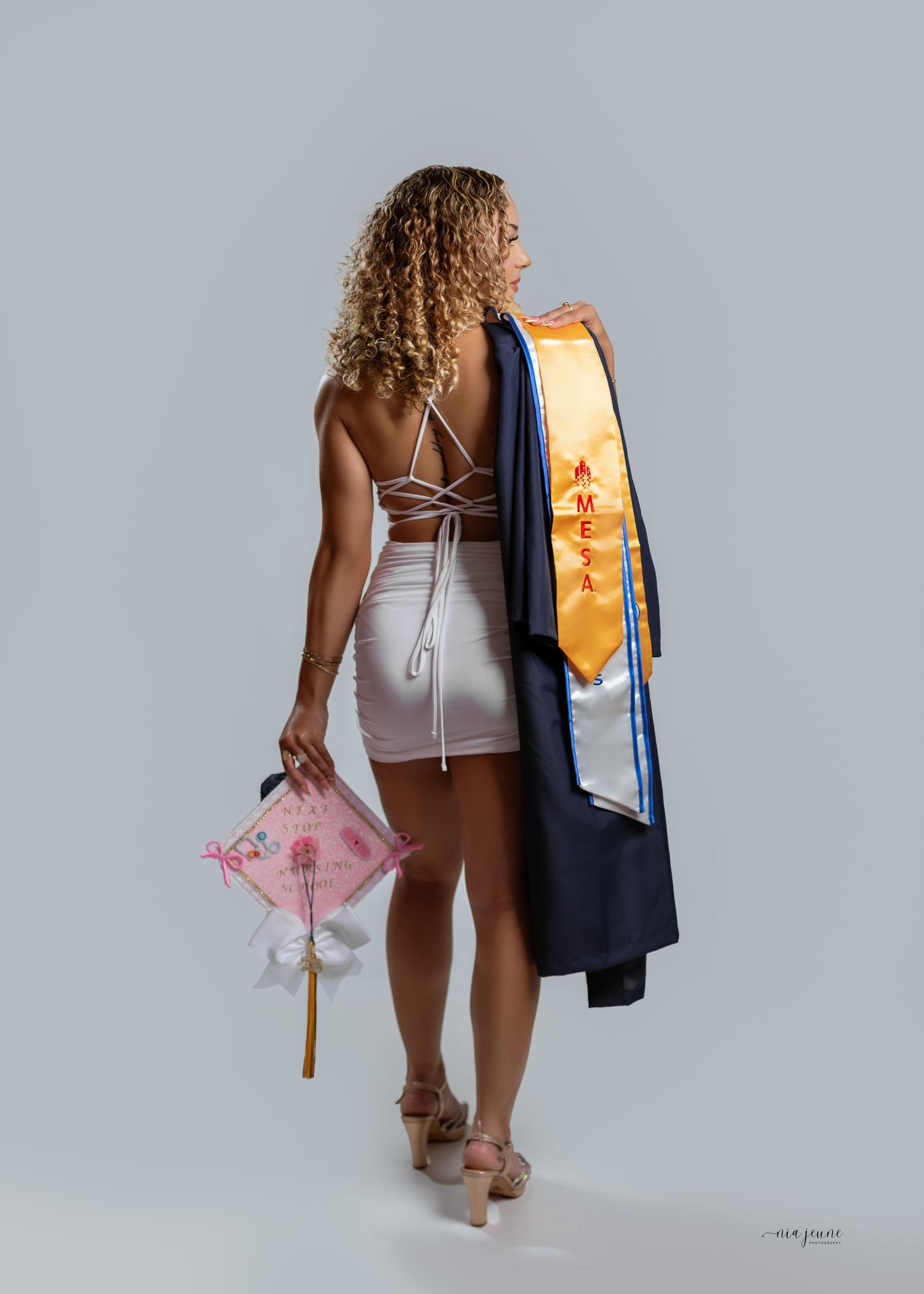Graduate in white dress holds decorated cap and MESA sash with navy gown draped over shoulder, posed from behind.