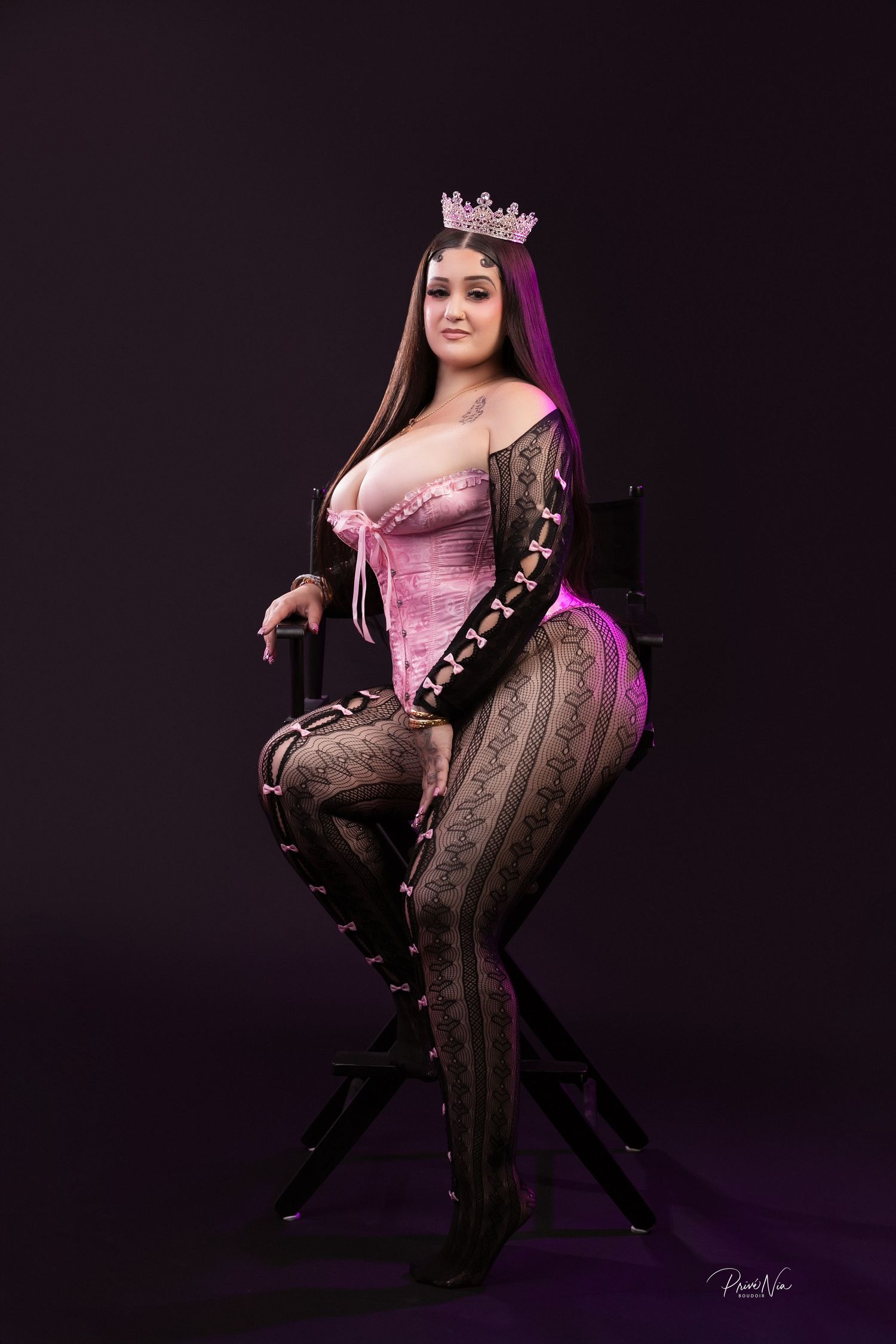 A curvy woman in a pink corset and lace leggings sits on a director's chair wearing a rhinestone crown.