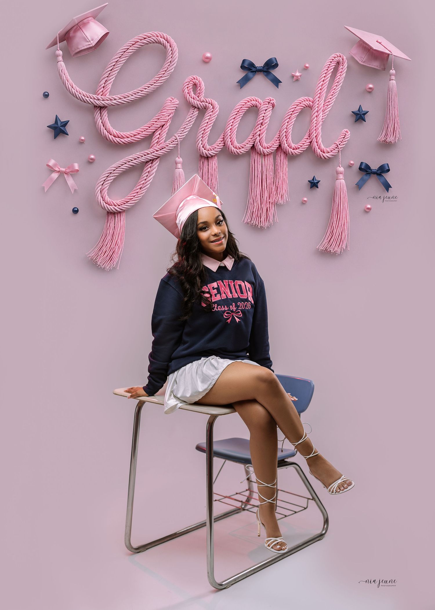 Smiling graduate in pink cap and senior hoodie sits on school chair under decorative pink Grad sign.