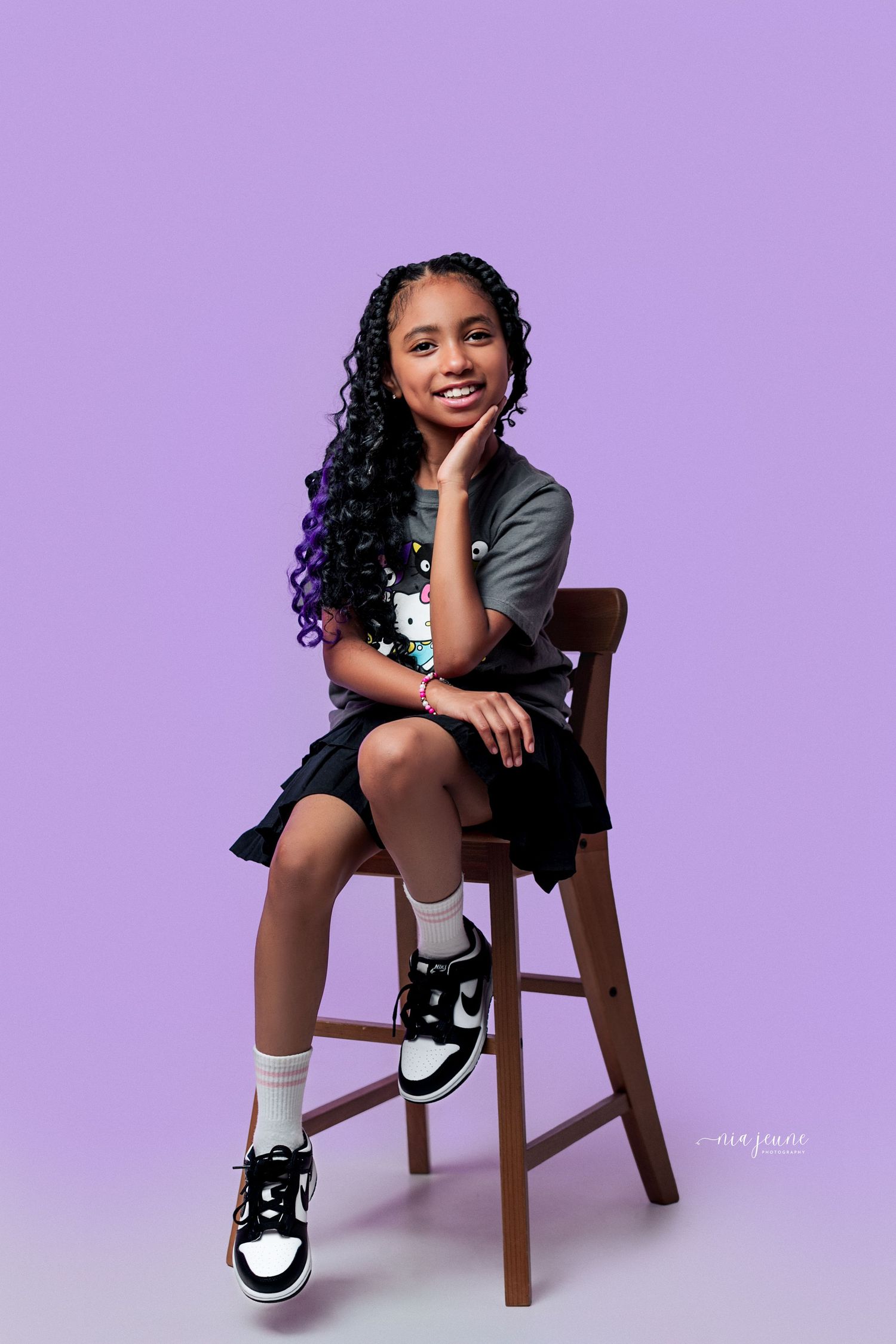 A young student in uniform sits on a wooden stool against a purple backdrop wearing black and white sneakers.