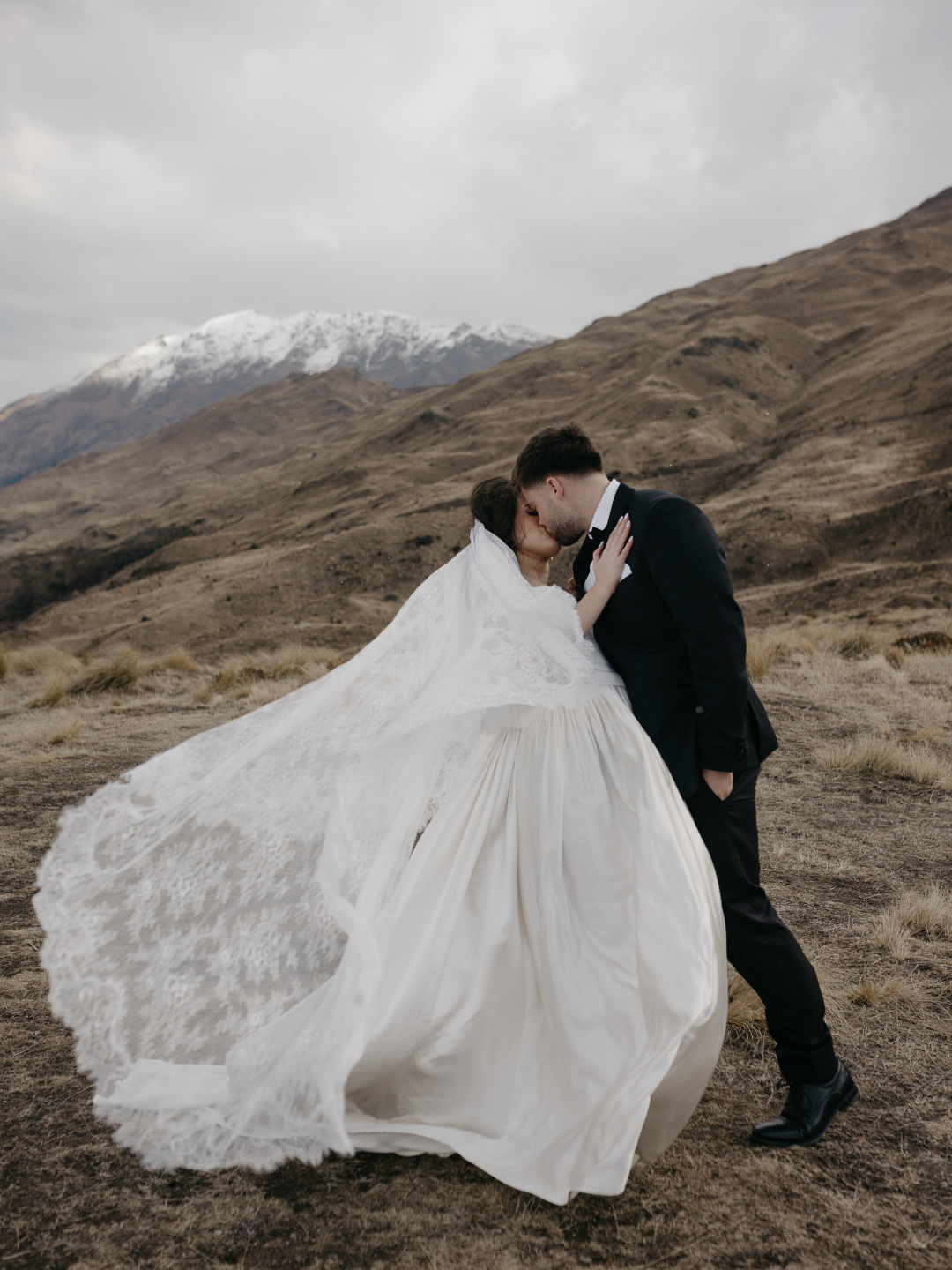 Bride and groom share a romantic kiss in front of snow-capped mountains, wedding veil flowing in the wind.