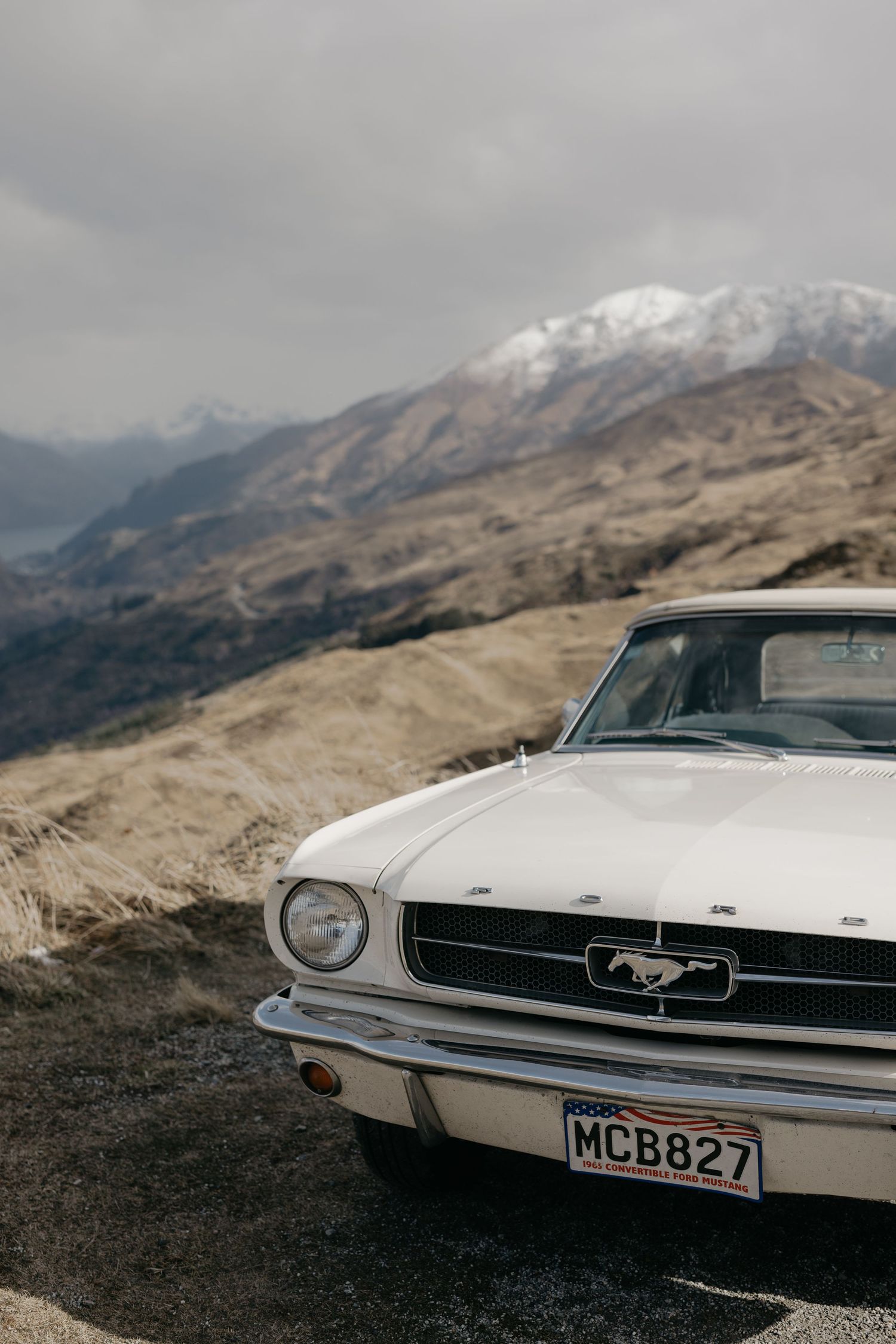 White vintage Ford Mustang parked on a gravel road with snow-capped mountains and overcast skies in the background.
