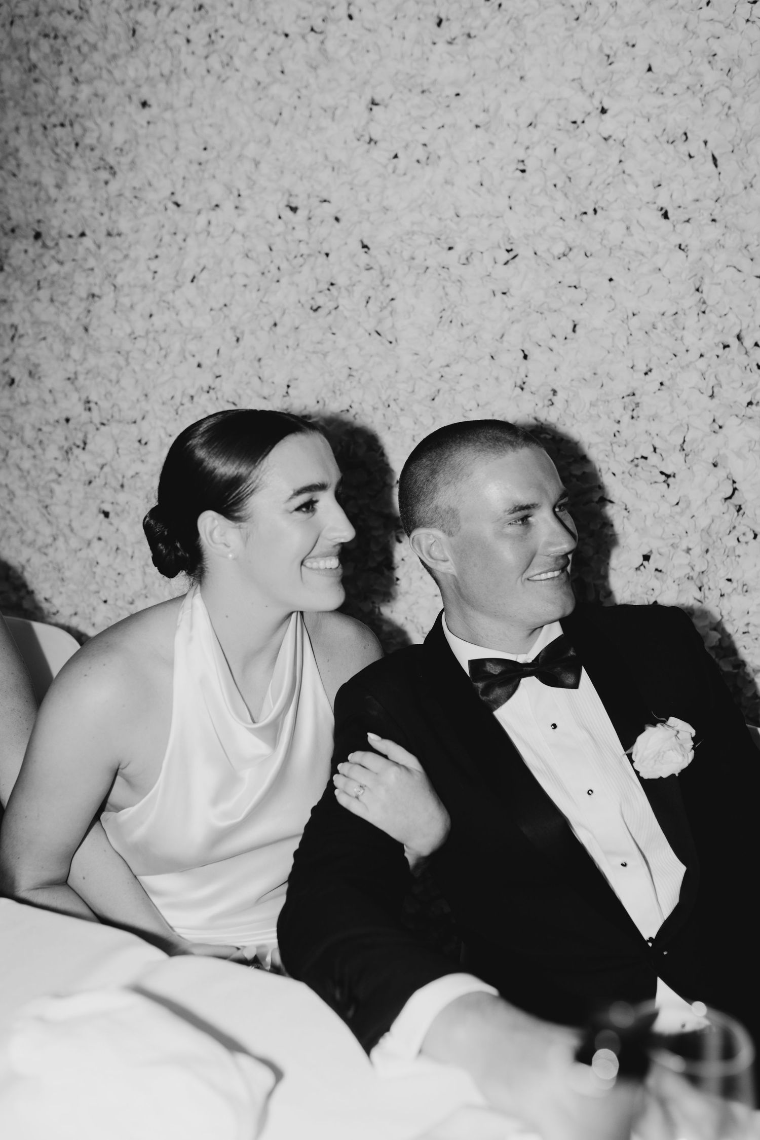 Black and white photo of a smiling bride and groom in formal attire seated together at their wedding reception.
