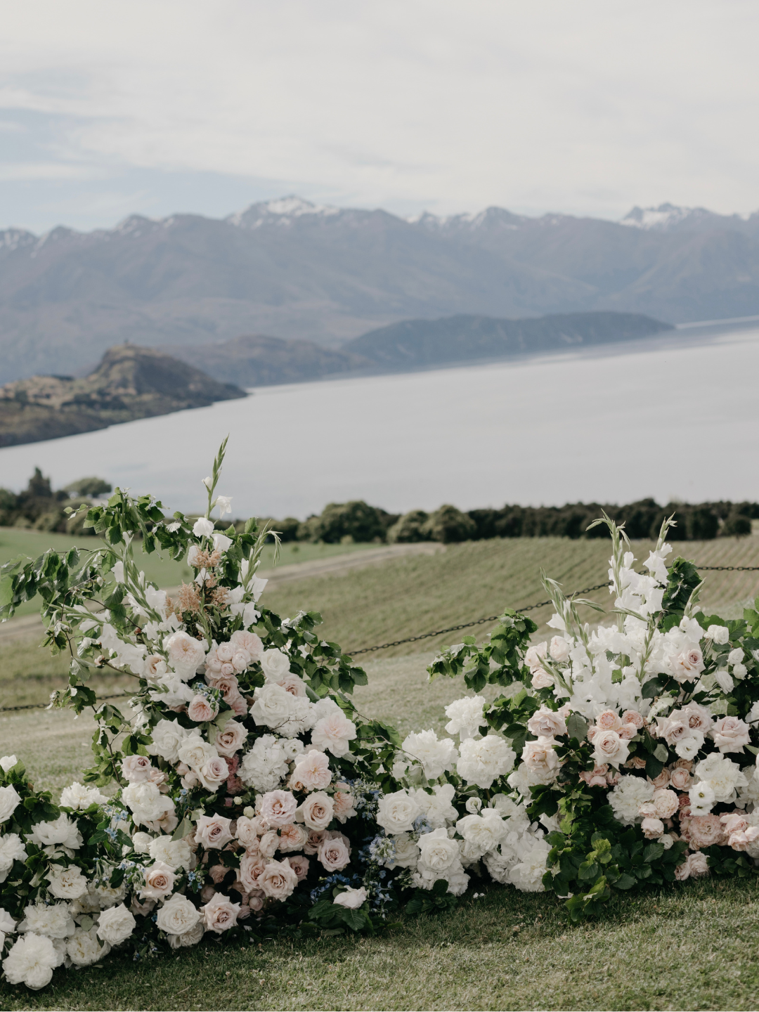 Elegant outdoor wedding arch adorned with blush and white roses overlooking a serene lake and snow-capped mountains in New Zealand.