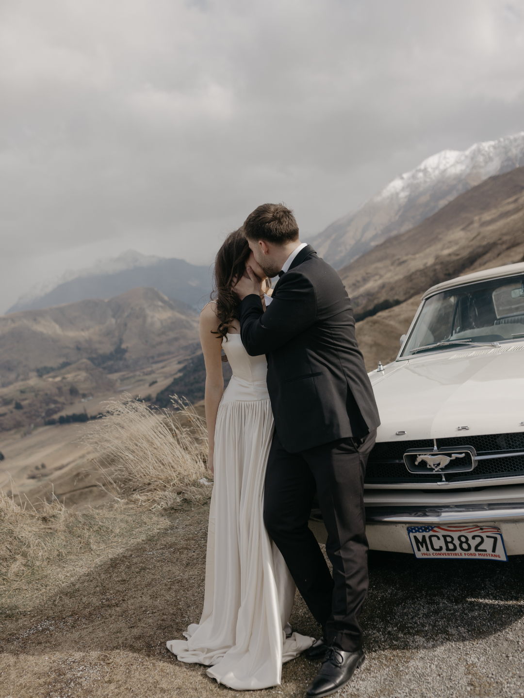 Couple sharing a romantic kiss in wedding attire beside a classic white Mustang with misty mountain backdrop in New Zealand.