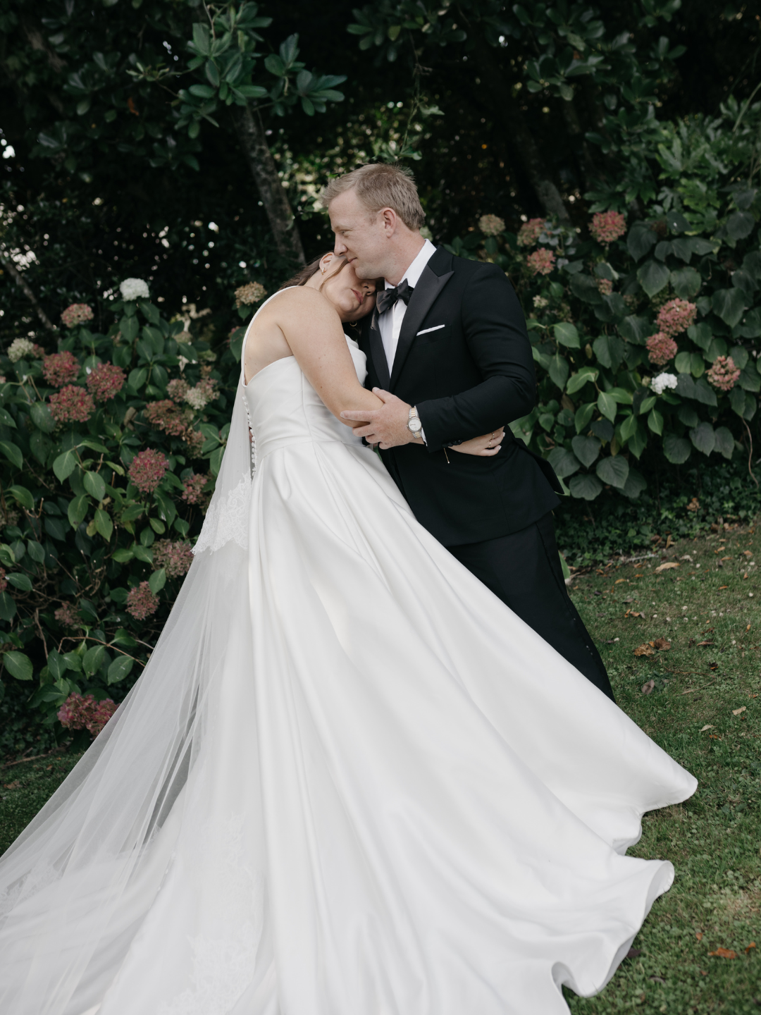 A bride and groom share a romantic kiss outdoors, surrounded by lush greenery and blooming flowers on their wedding day.