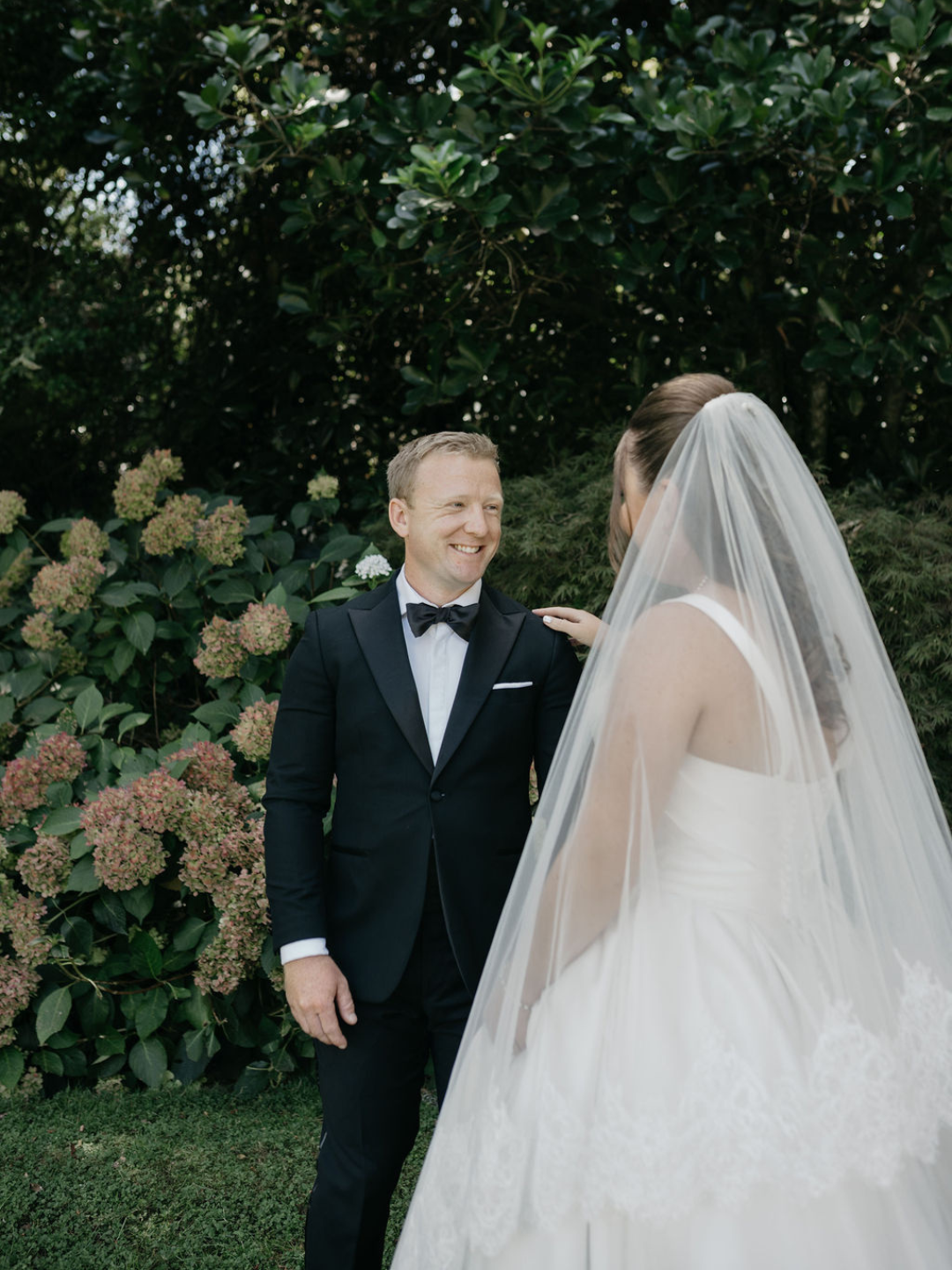 Groom in black tuxedo smiling at bride in white wedding dress and veil, standing outdoors near blooming hydrangeas.