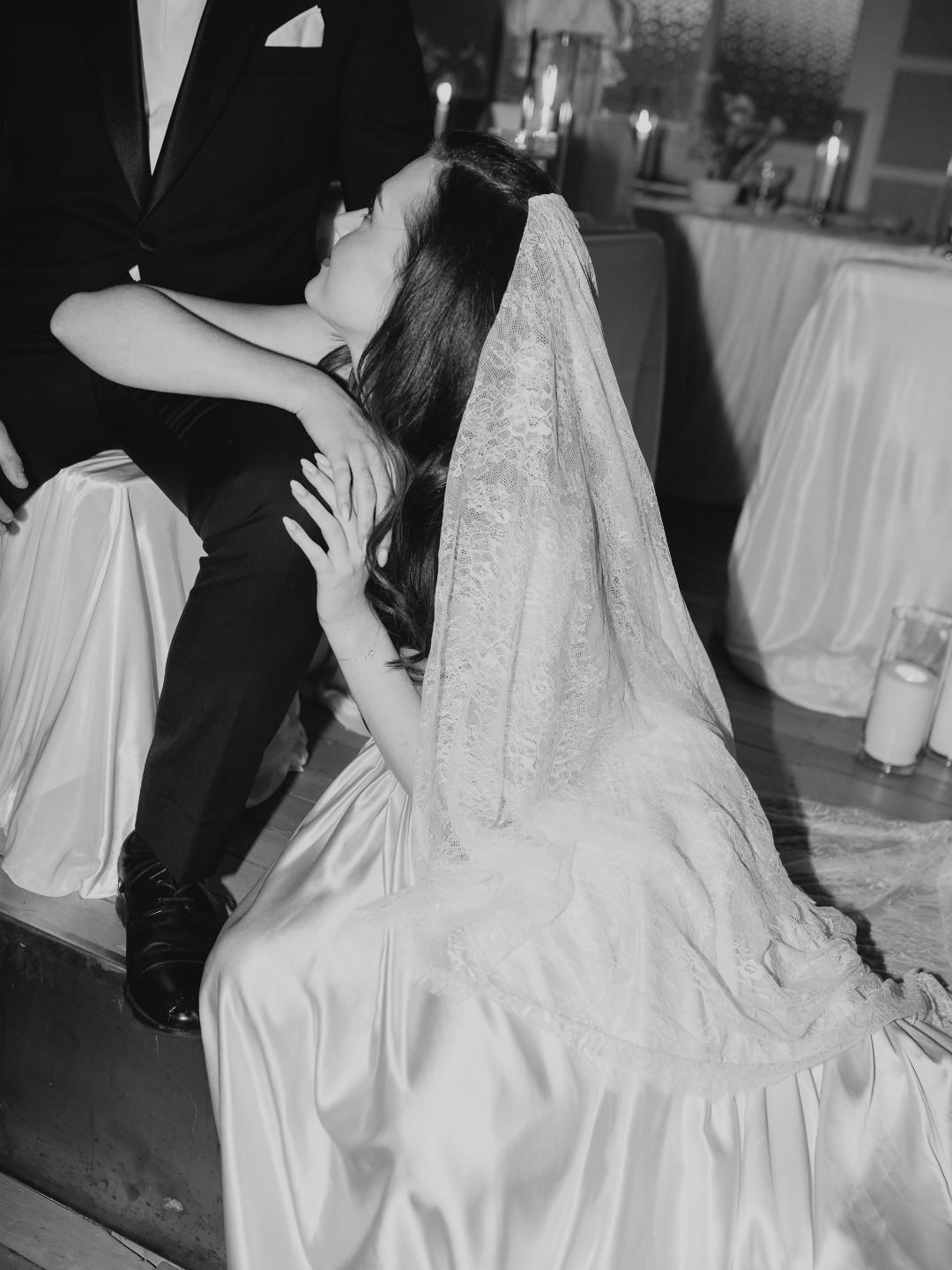 Black and white photo of bride in satin gown and veil sharing a romantic kiss with groom in tuxedo at wedding reception.