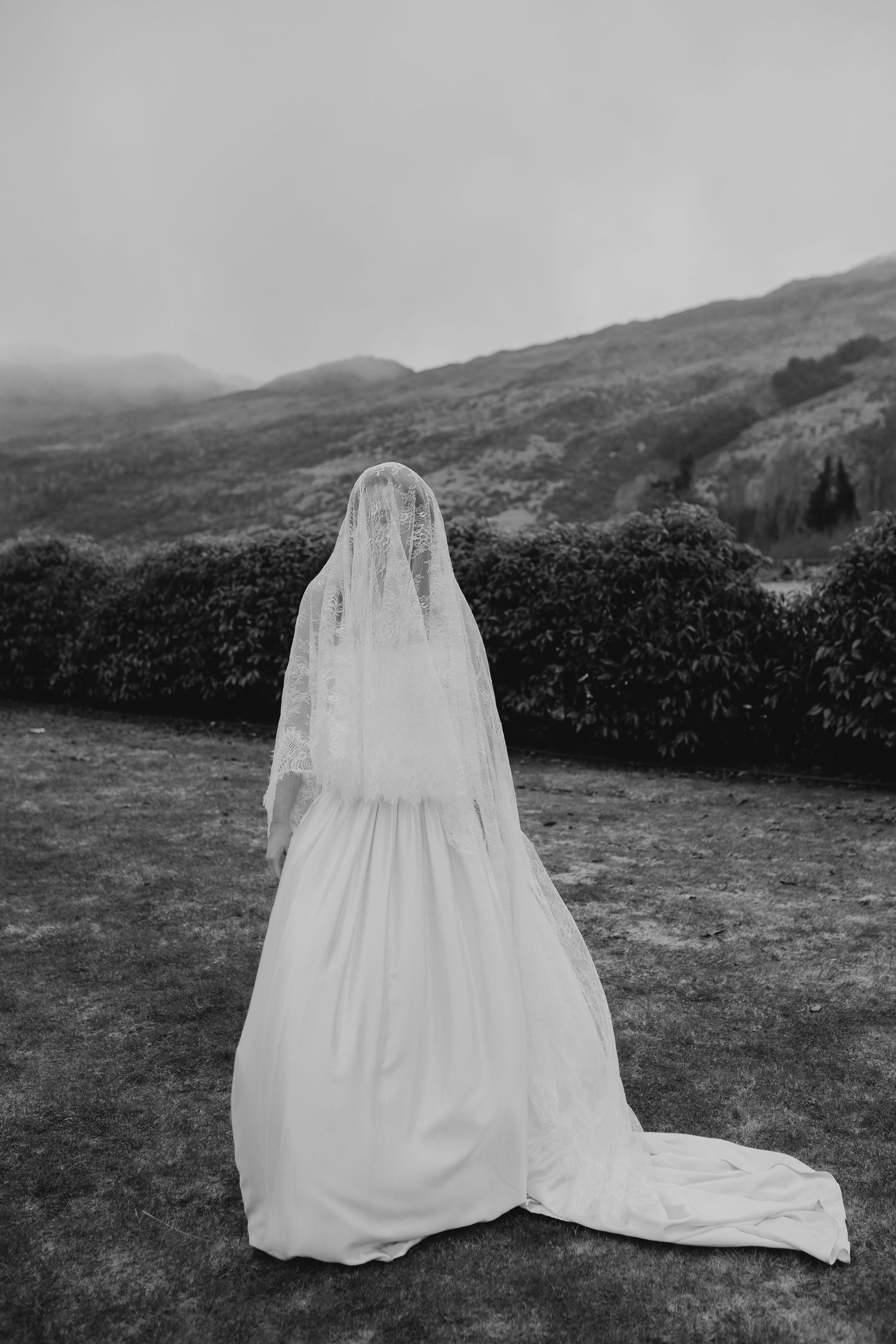 Bride in white gown and lace veil stands facing misty mountain landscape in elegant black and white photo.