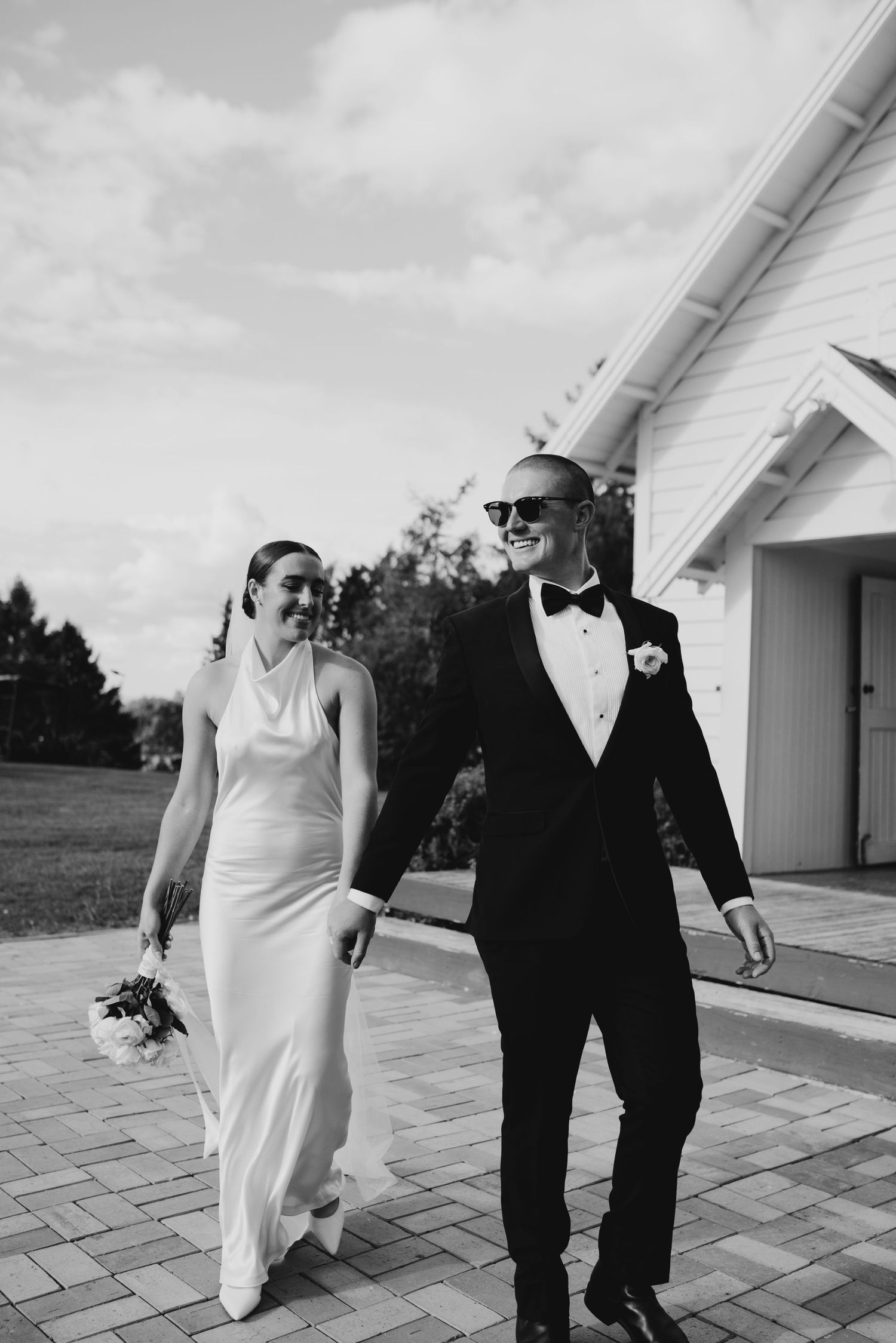 A bride and groom walking together outside a white chapel, the groom is wearing sunglasses and tuxedo, and the bride is in an elegant gown.