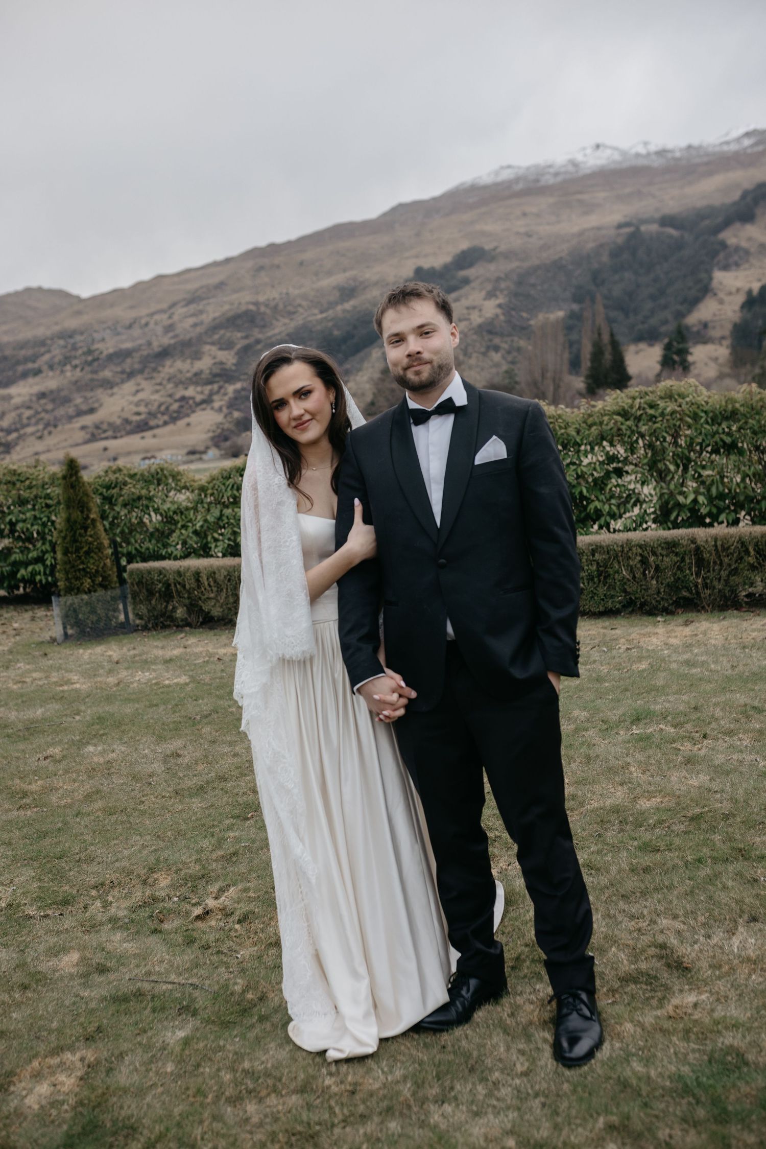 A bride and groom pose together outdoors on their wedding day with a scenic mountain backdrop in New Zealand.