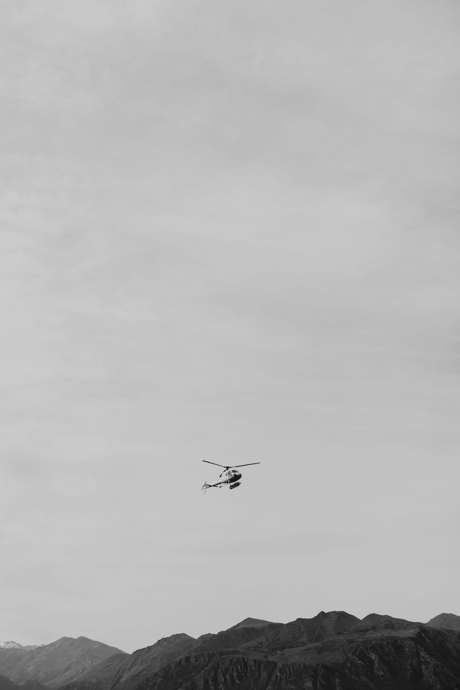 Black and white photo of a helicopter flying over mountain peaks against an overcast sky.
