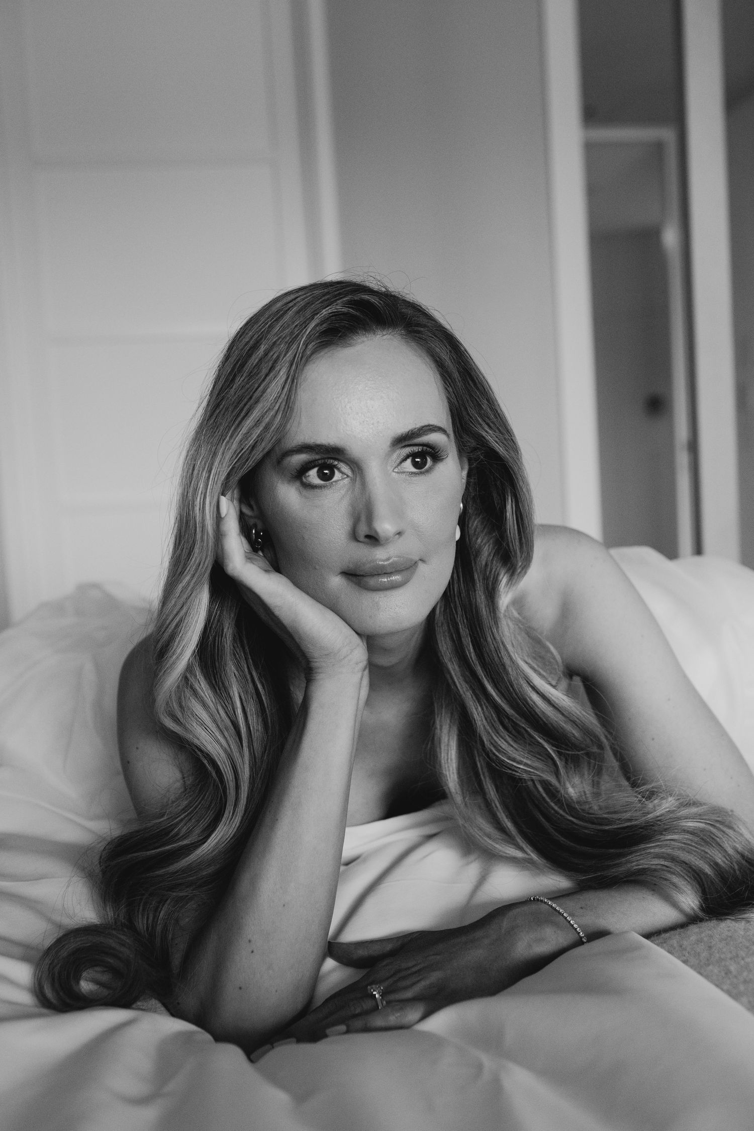 Black and white portrait of a woman with long wavy hair resting her chin on her hand while lying on a bed.