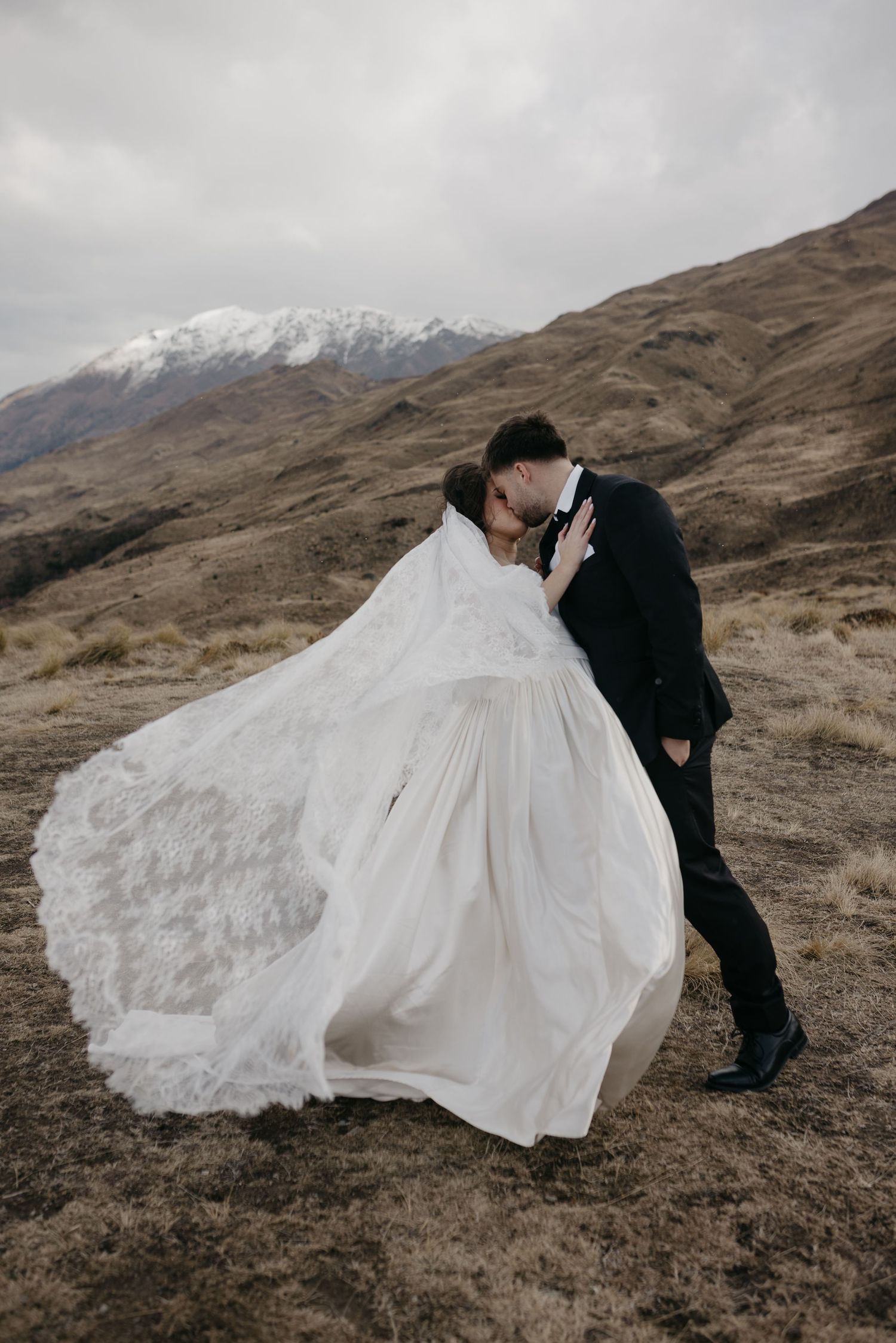 Bride and groom share a romantic kiss on a rugged mountainside, her lace wedding dress flowing in the wind.