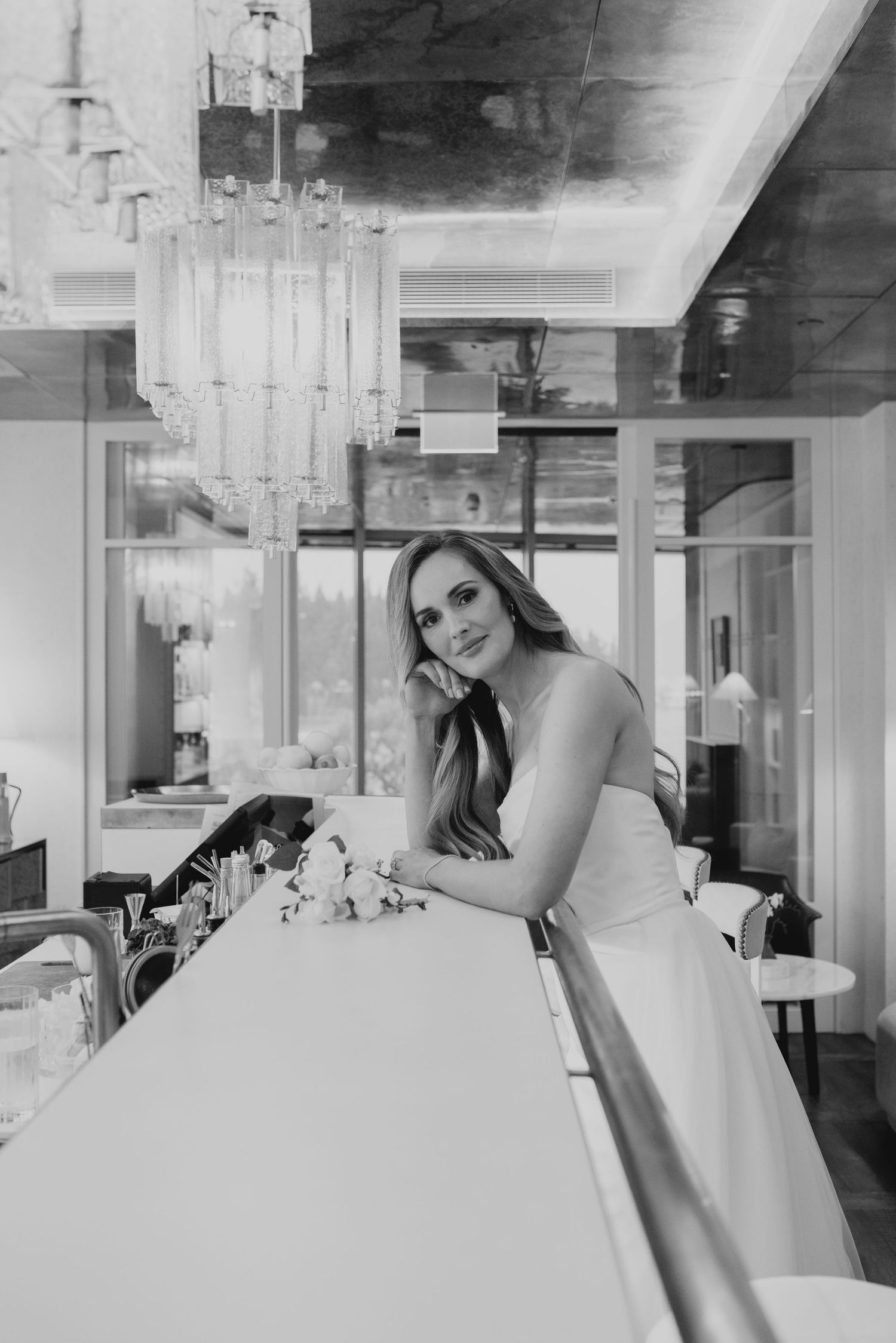 Black and white photo of an elegant bride leaning on a bar counter in a luxurious venue with chandelier.