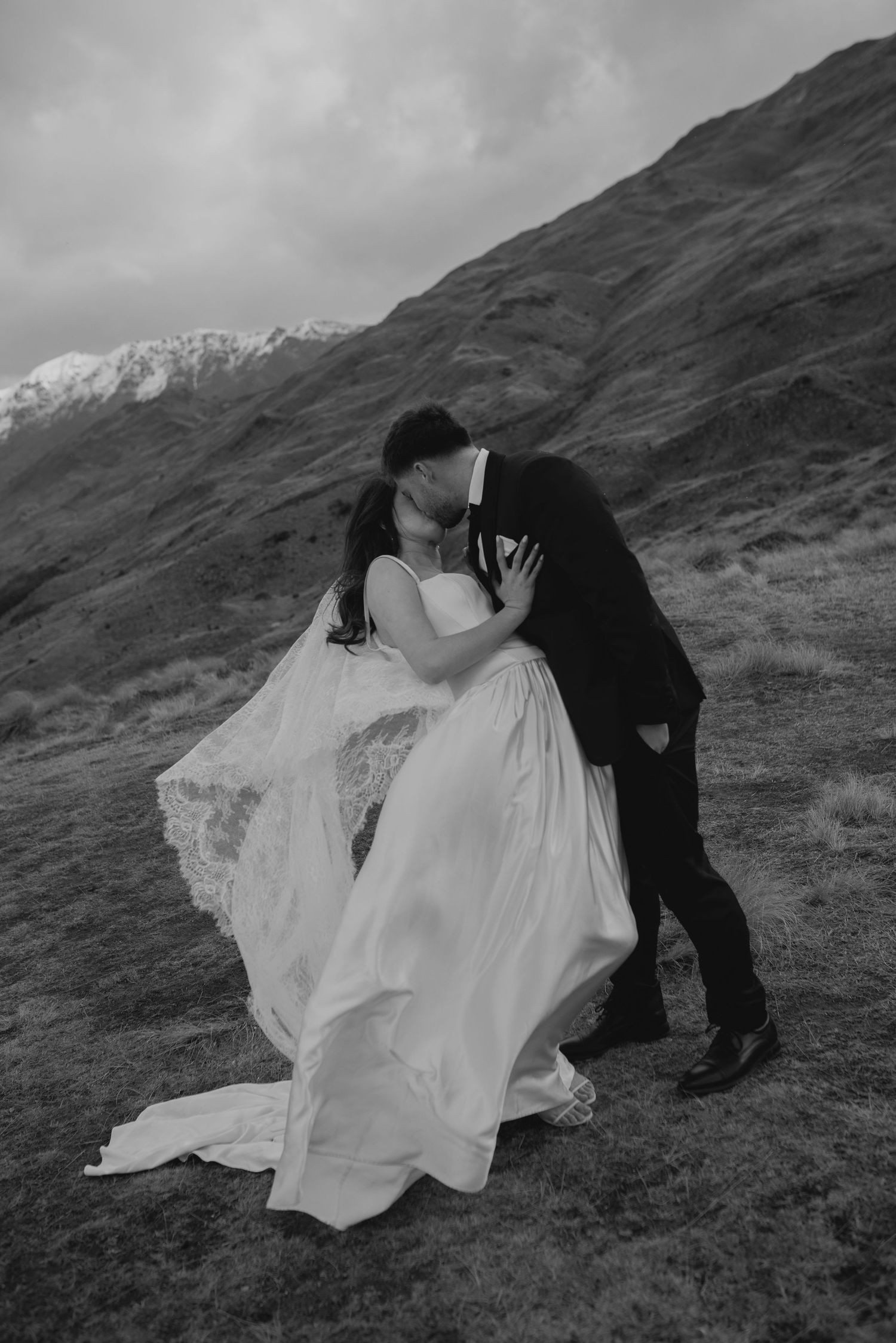 Black and white photo of a couple kissing in wedding attire on a mountain hillside with snow-capped peaks behind them.