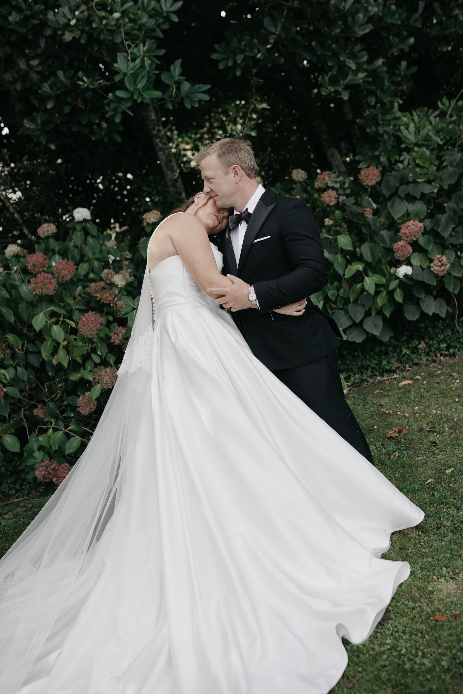 A bride and groom share a romantic kiss in a lush garden, her white gown flowing gracefully as he holds her close.