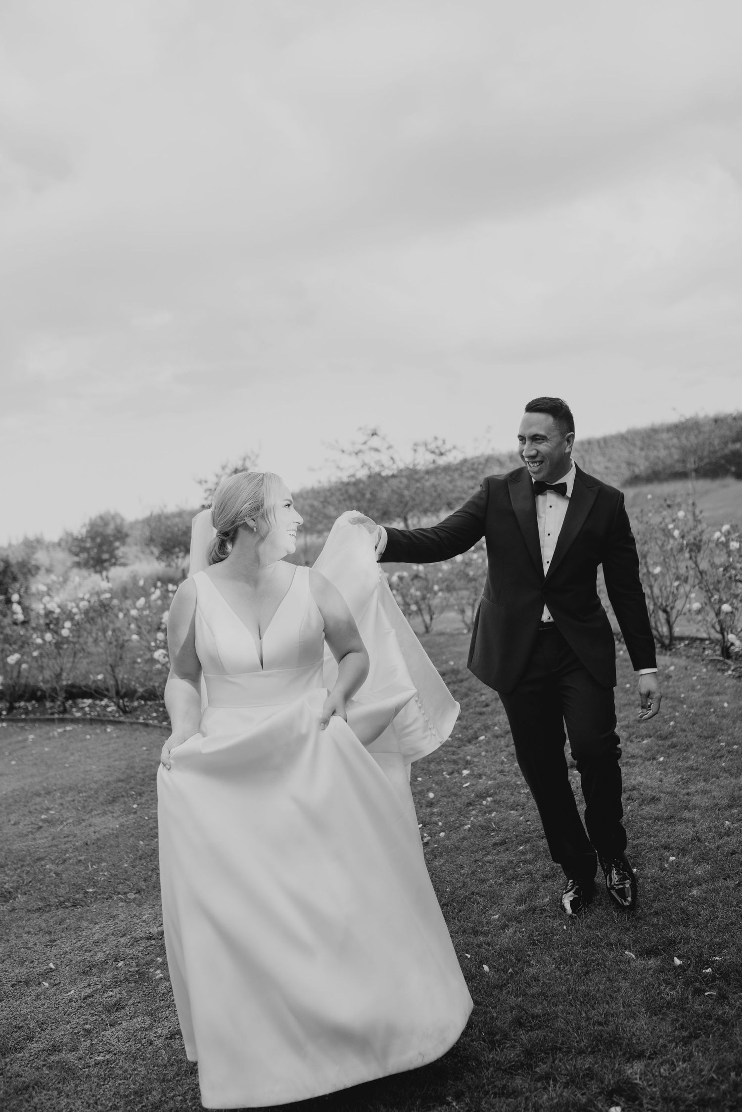 Black and white photo of a smiling bride in white gown and groom in tuxedo walking together outdoors.