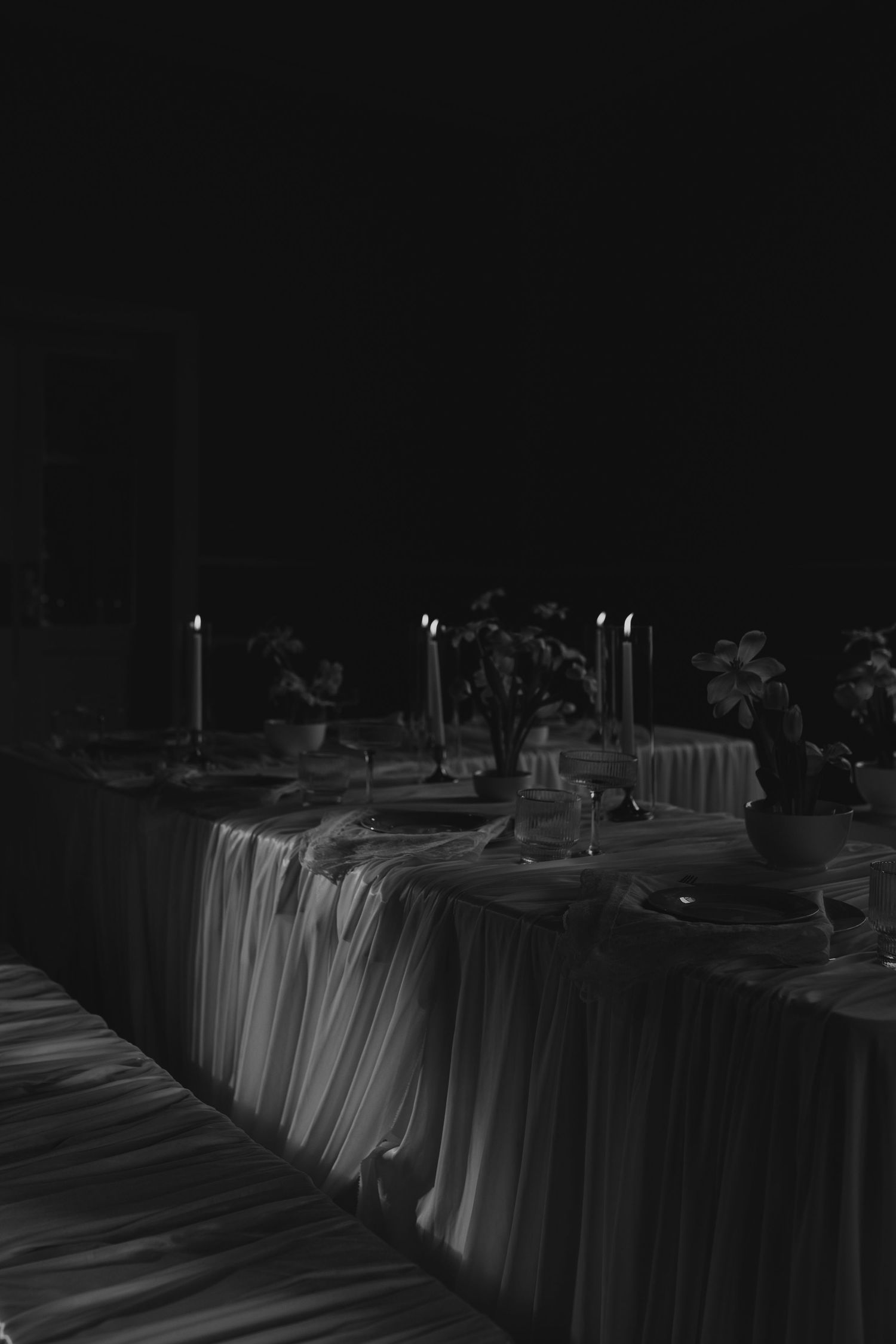 Black and white photo of an elegantly set dining table with lit candles and floral centerpieces in a dimly lit room.