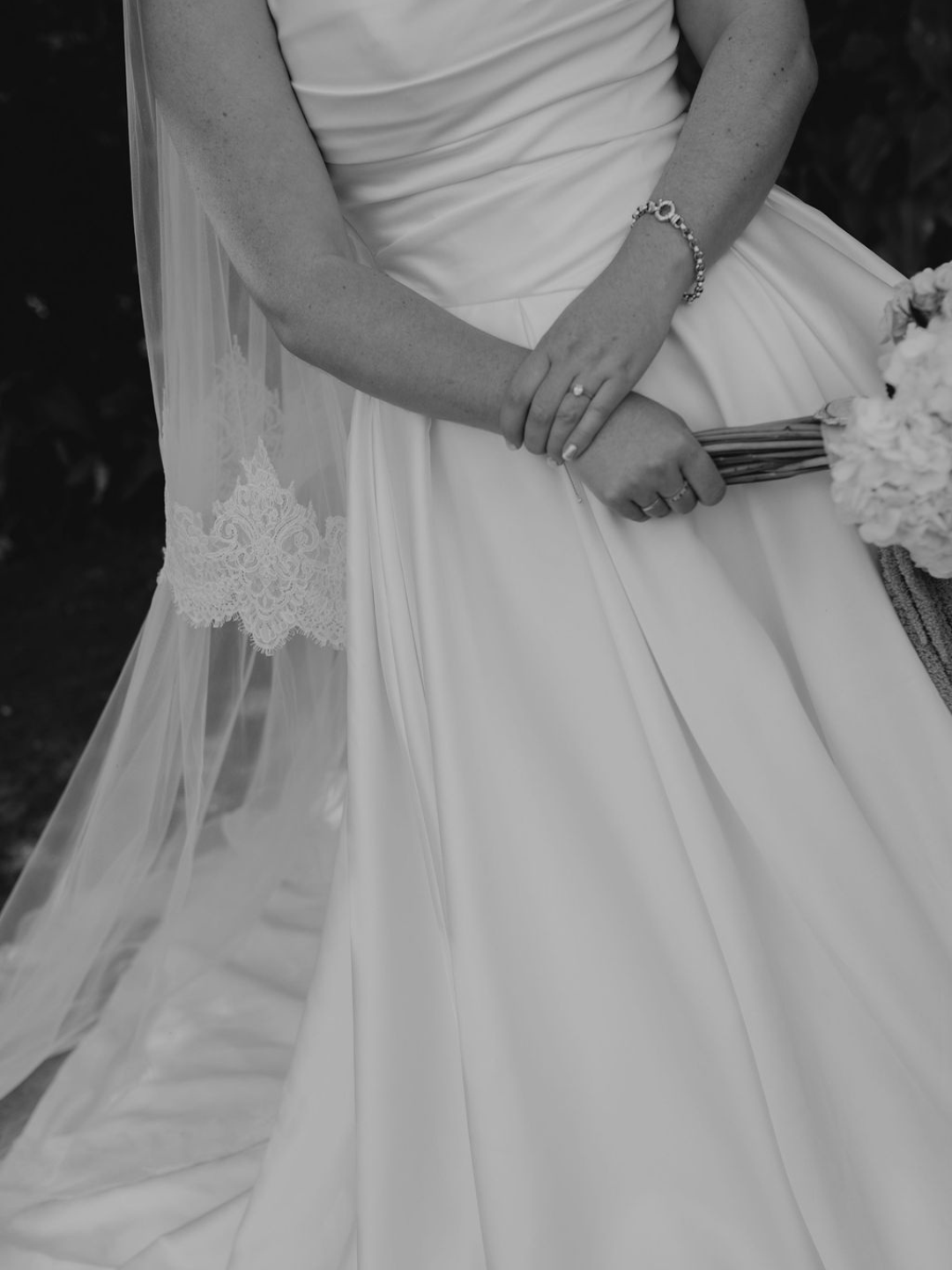 Black and white photo of a bride in a white gown holding a bouquet, showcasing her lace veil and diamond bracelet.