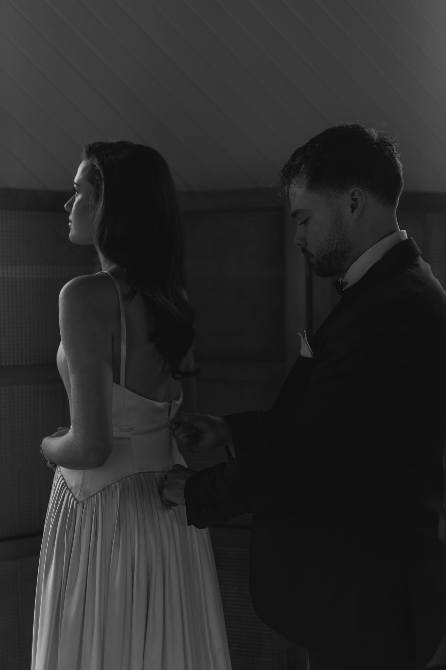 Black and white photo of a groom fastening the back of a brides white dress in a dimly lit room.