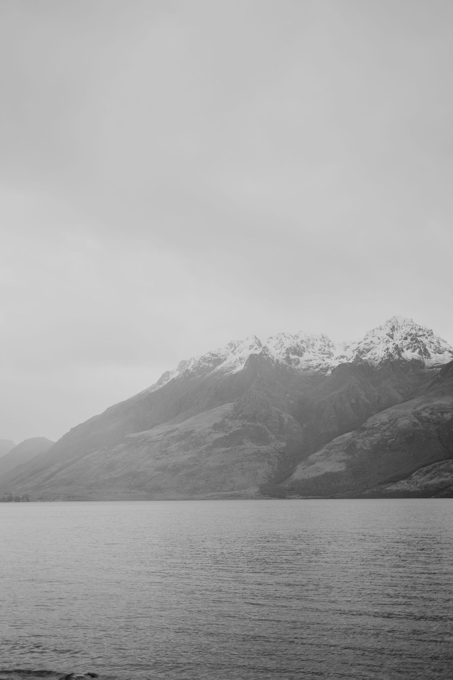 Black and white landscape of snow-capped mountains reflected in a calm lake under an overcast sky.