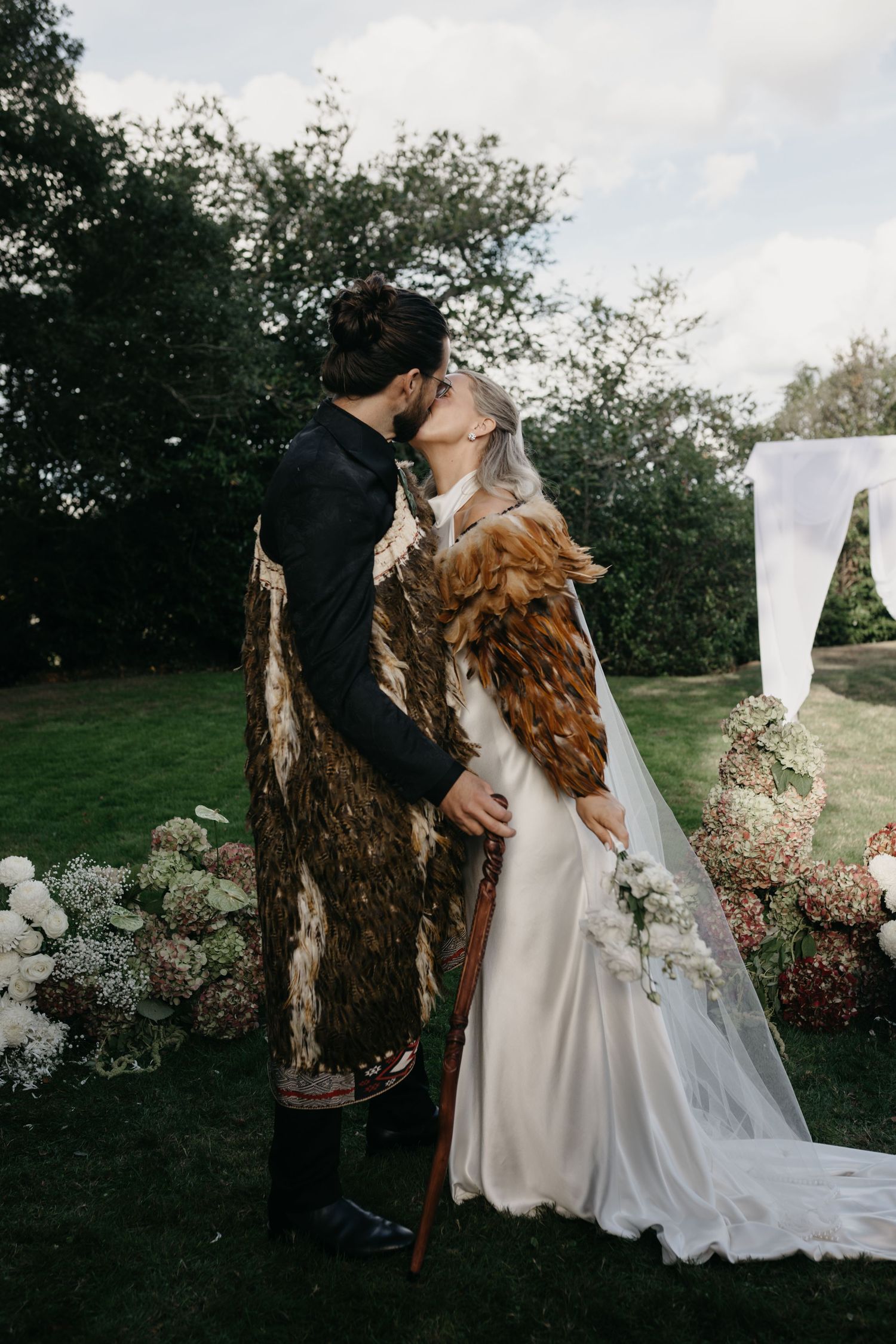 Couple sharing first kiss at outdoor wedding ceremony, bride in white gown with veil, groom in feathered cape, floral arrangements.