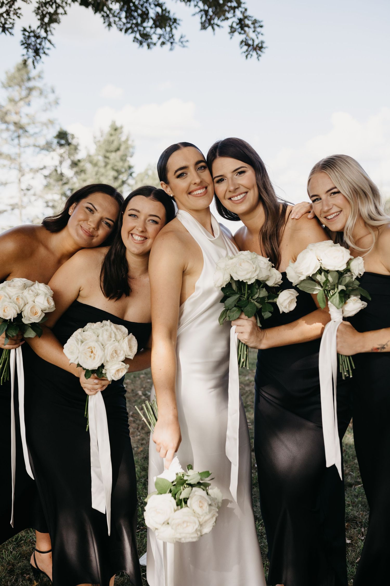 Bride in white satin gown poses with four bridesmaids in black dresses holding white rose bouquets outdoors.