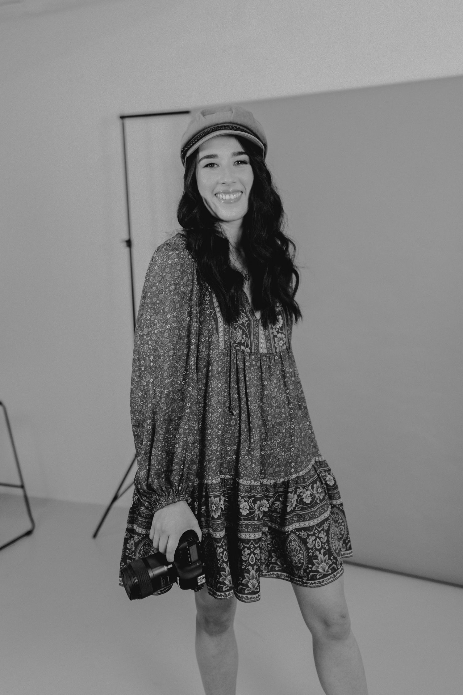 Smiling woman in floral dress and hat holding a camera in a photography studio, black and white photo.