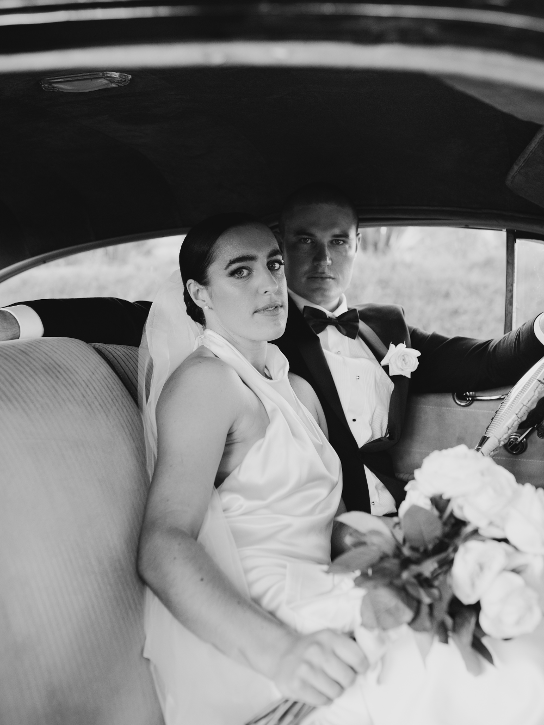 Black and white photo of a bride holding bouquet and groom in tuxedo sitting together in a vintage car.