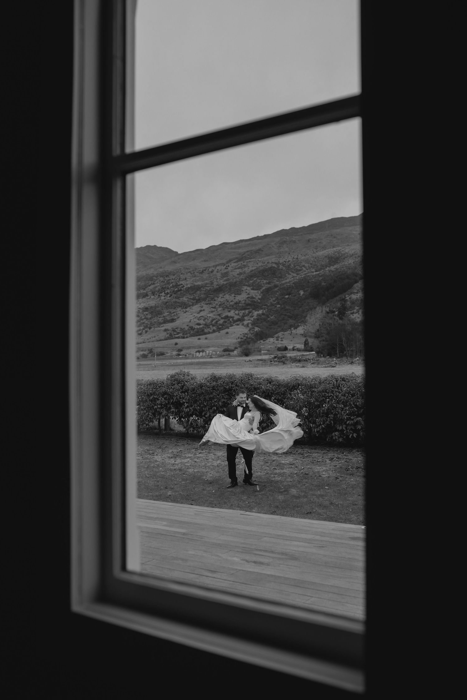 Black and white photo of bride and groom embracing outdoors, veil flowing in wind, scenic mountain landscape viewed through window.