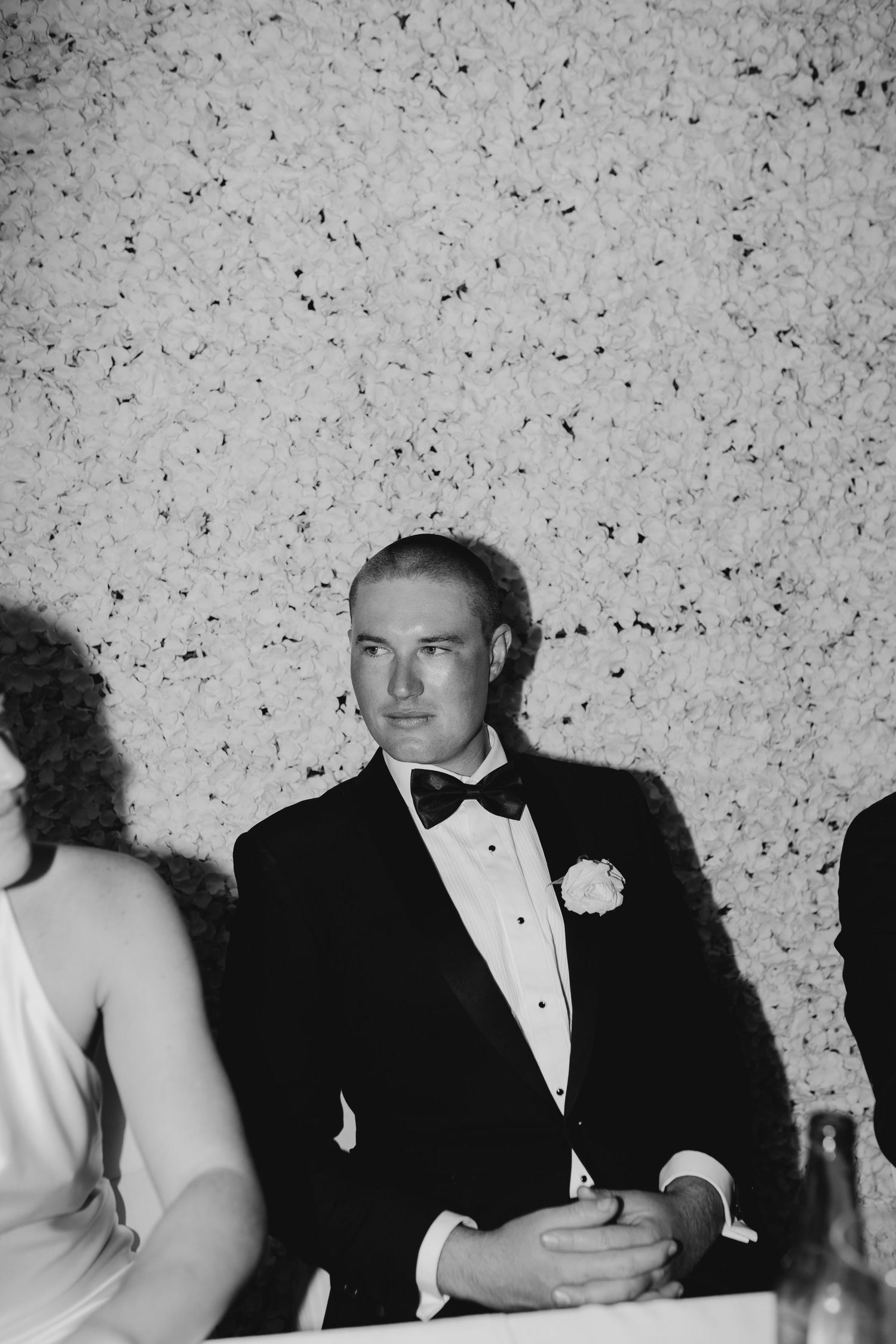 Black and white photo of a groom in a tuxedo with bow tie and boutonniere seated at a wedding reception.