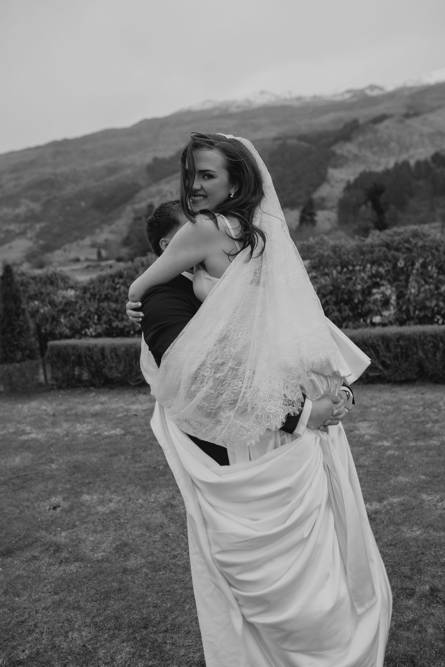 Black and white photo of a bride in a veil being lifted by her groom outdoors with mountains in the background.
