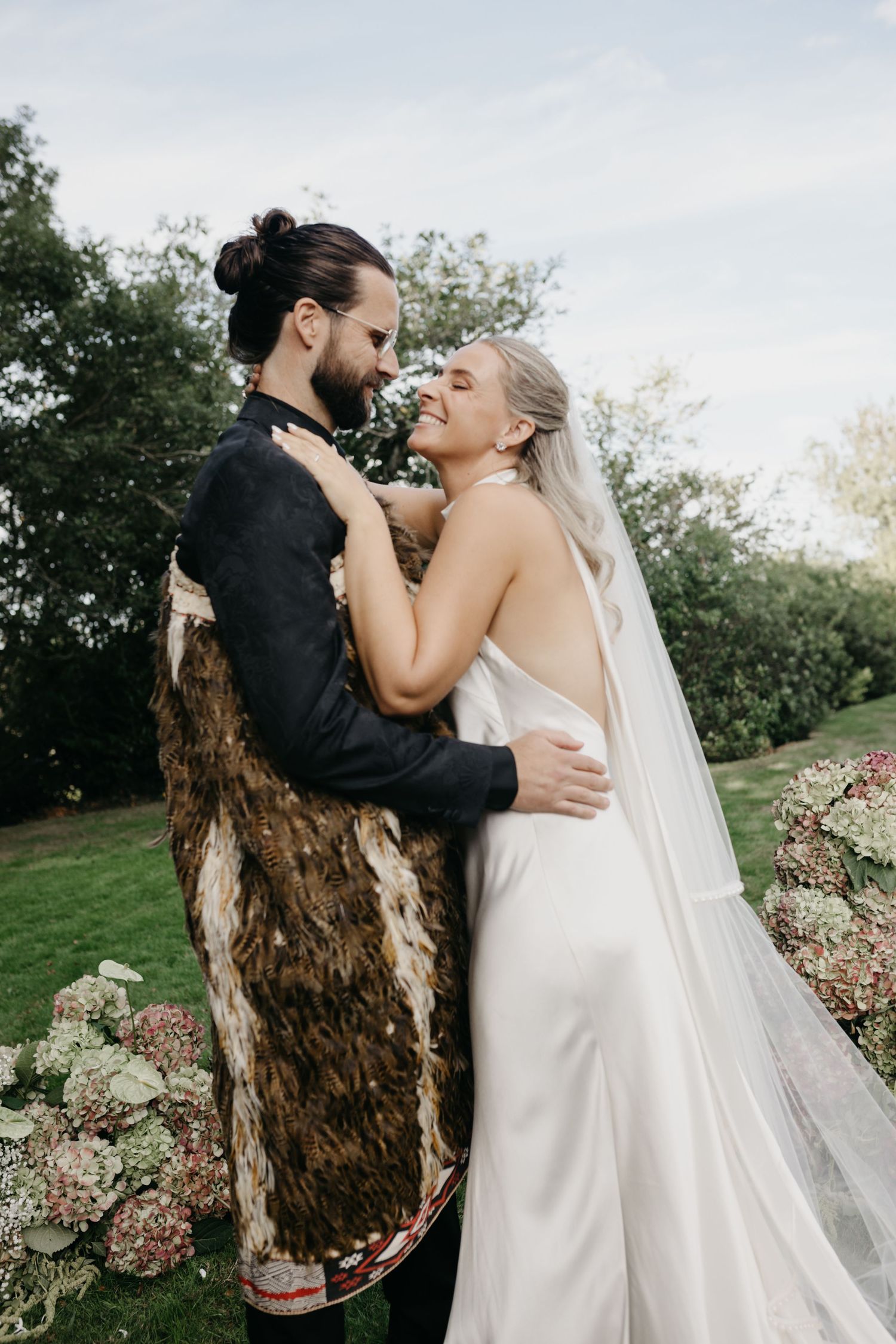 Bride in white satin gown and veil embraces groom in traditional Maori cloak, laughing together outdoors at wedding.