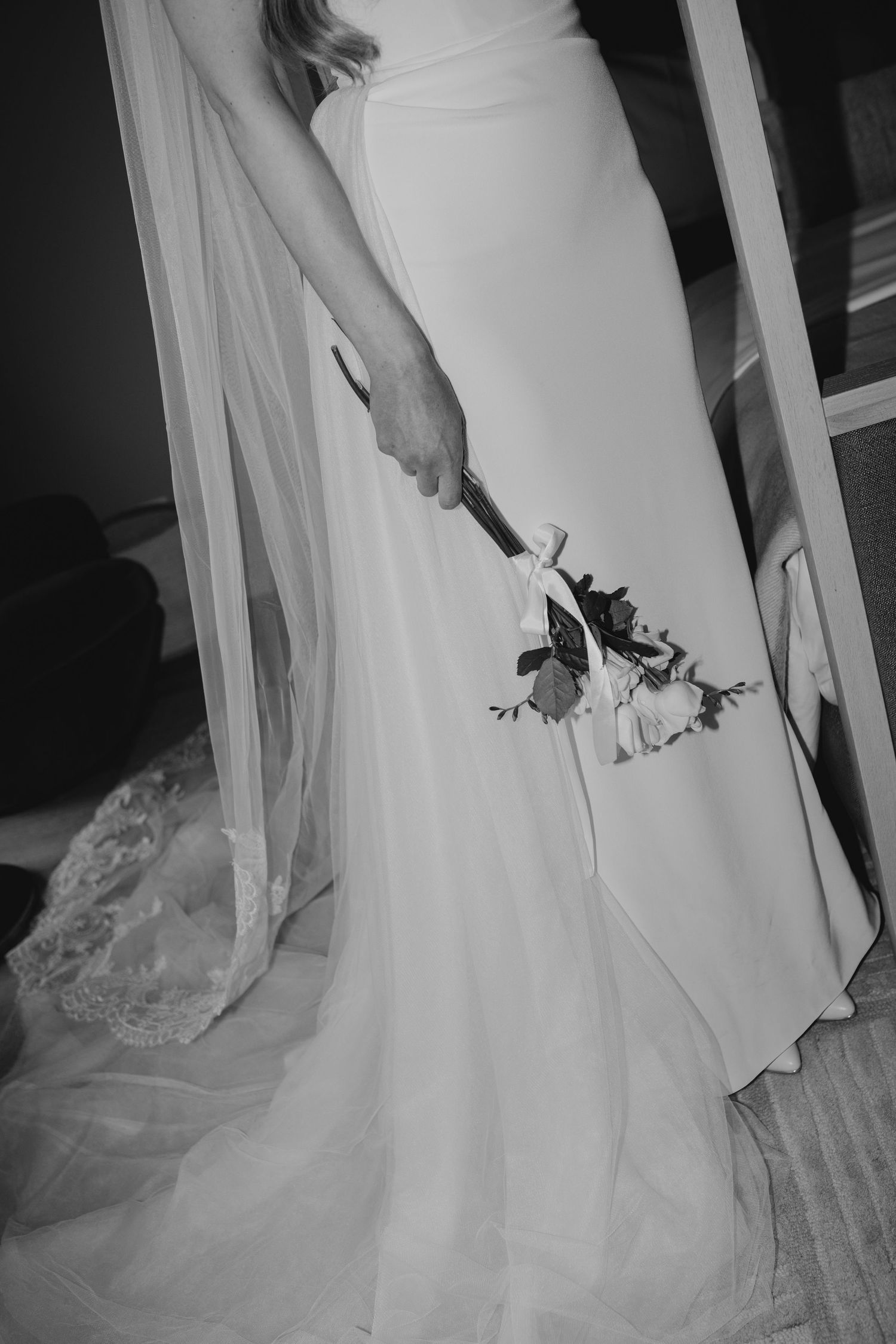 Black and white photo of a bride in a white gown holding a small floral bouquet with a lace veil trailing beside her.