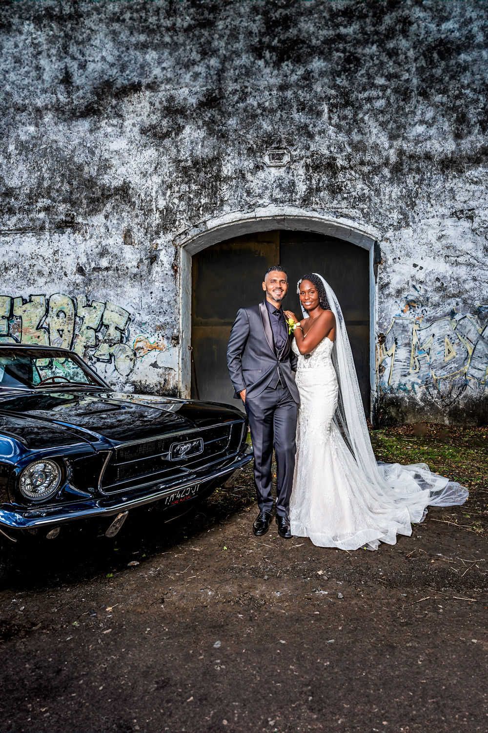 Un couple de jeunes mariés pose en tenue de soirée à côté d'une Ford Mustang vintage contre un mur de béton patiné avec des graffitis.