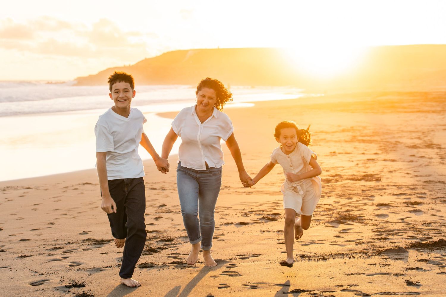 Family of three running joyfully on a beach at golden sunset, holding hands and smiling.