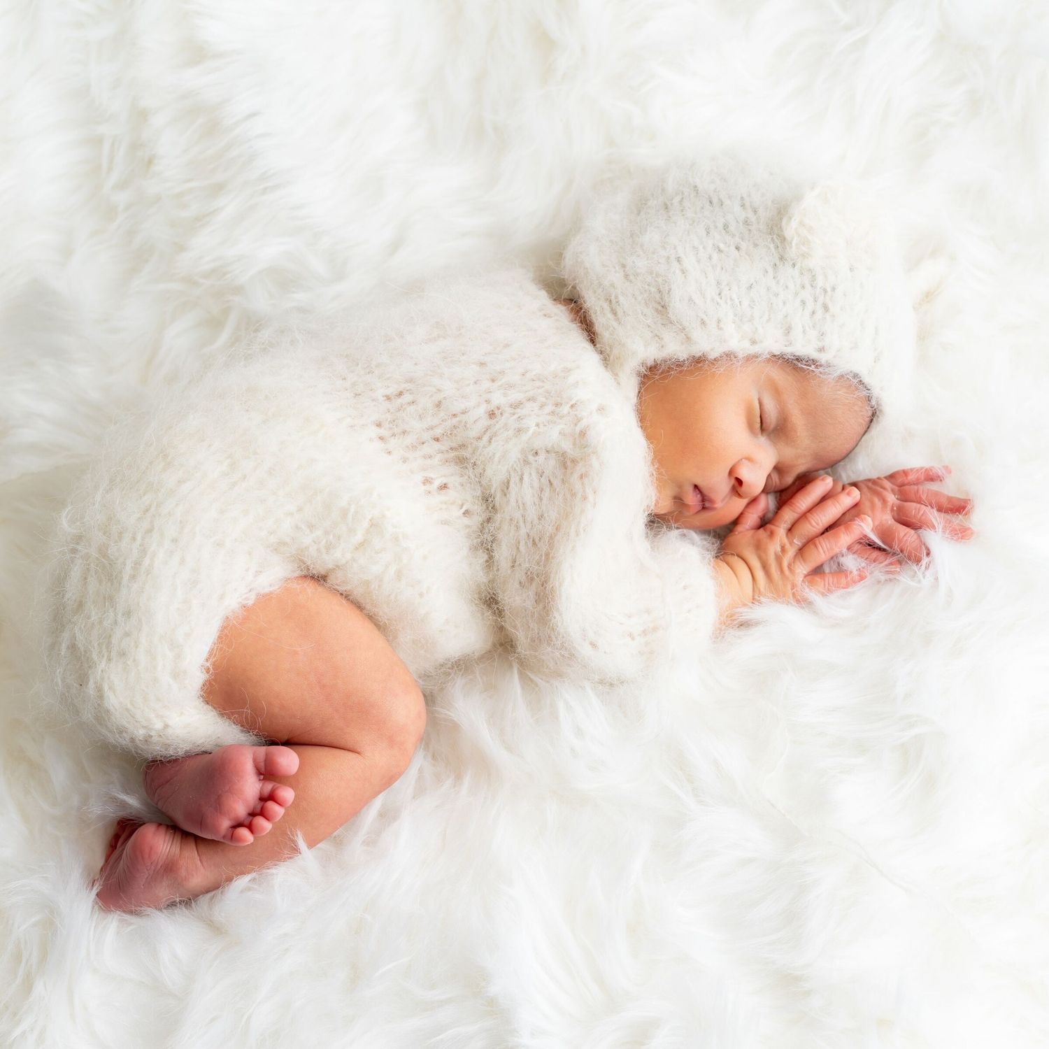 Newborn baby sleeping peacefully on white fur blanket wearing a cozy knit bear outfit and hat.
