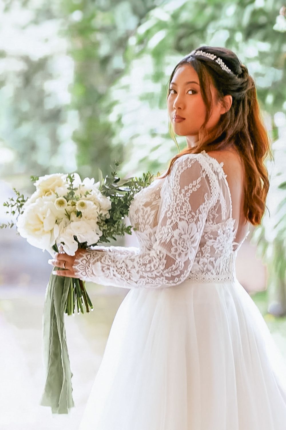 A bride in a long-sleeved lace wedding dress holds a white bouquet while looking over her shoulder in a dreamy outdoor setting.