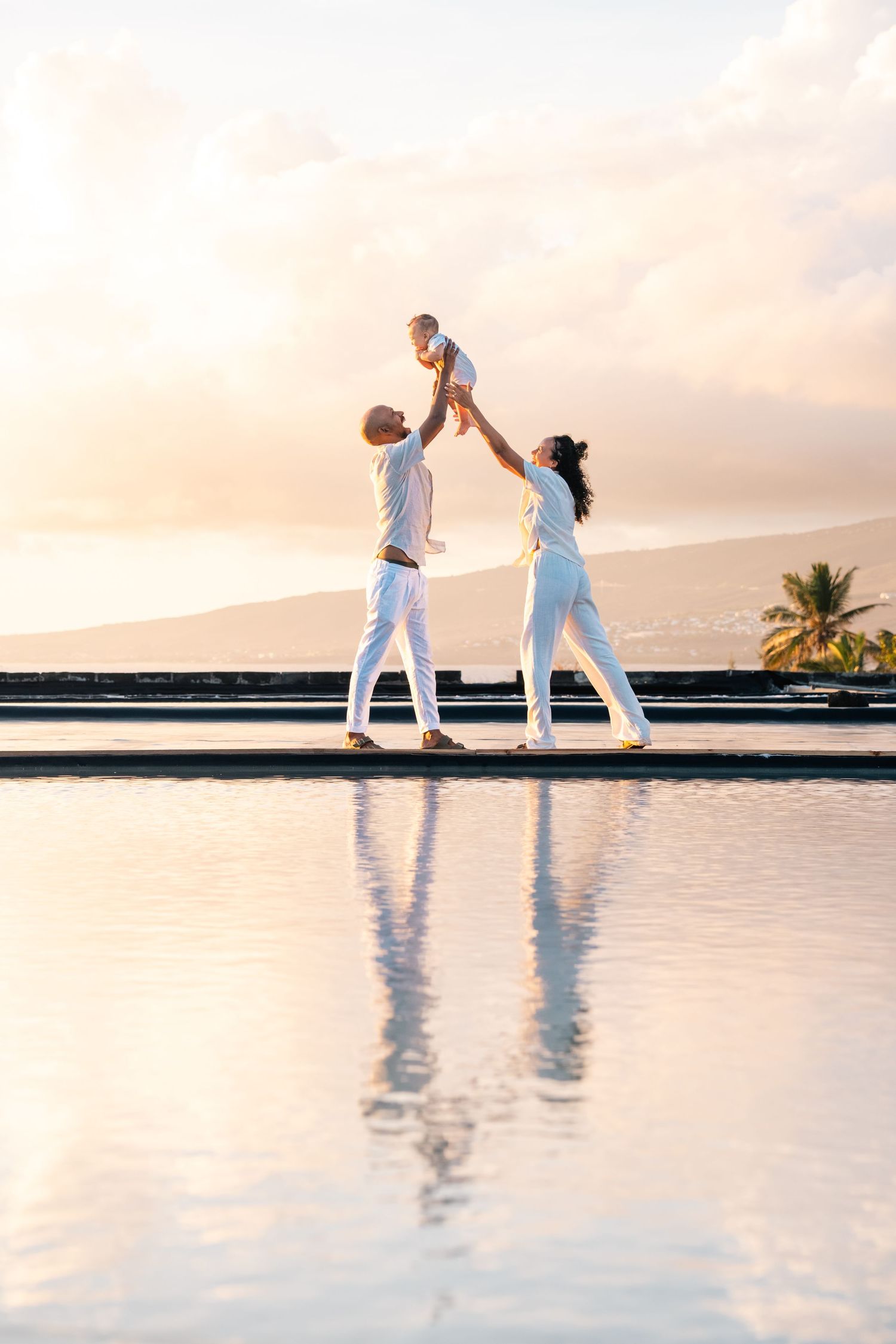Happy family in white outfits lifting baby by infinity pool at sunset with ocean and mountains backdrop.