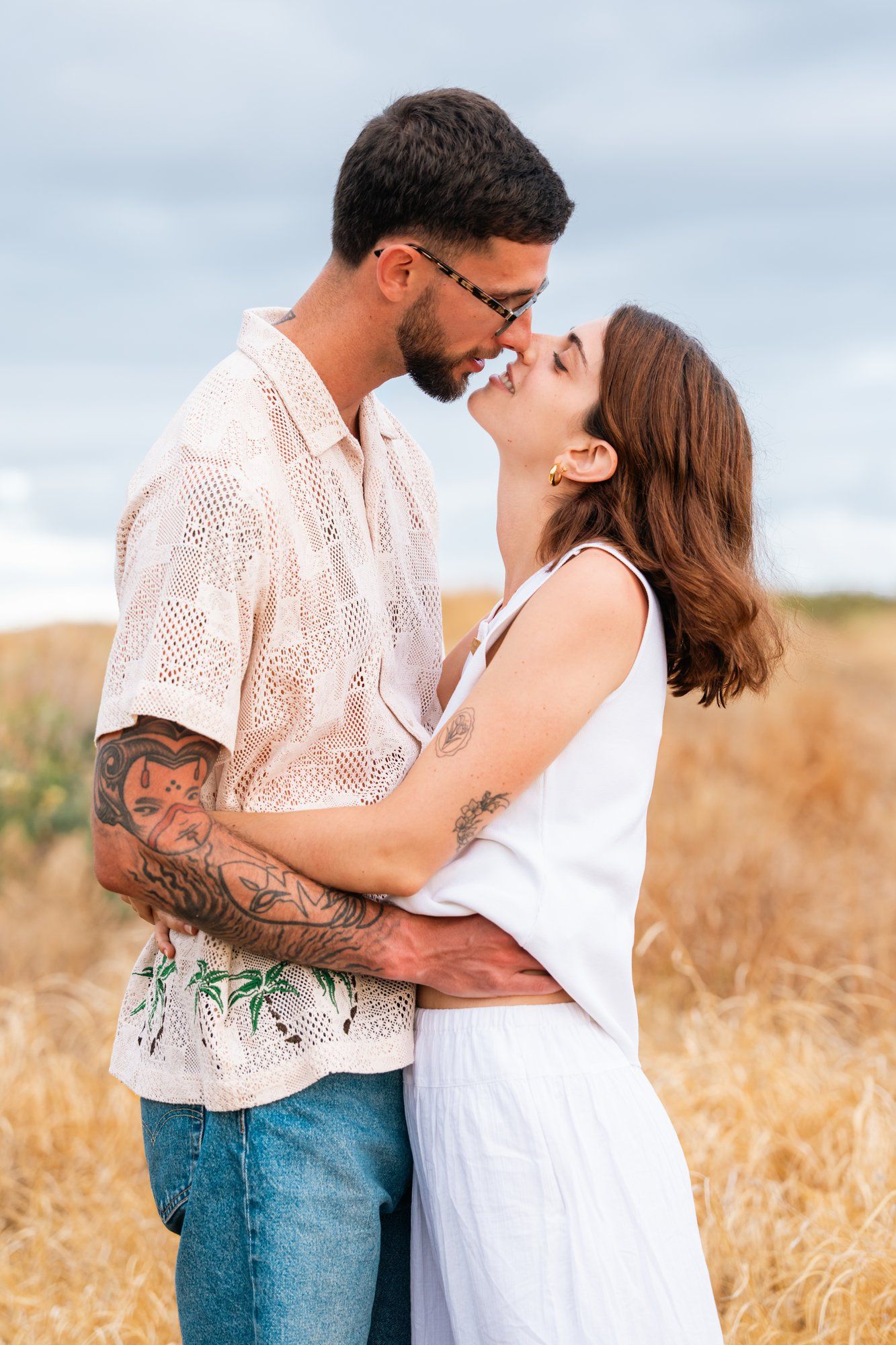 Romantic couple embracing and about to kiss in a golden field, man with tattoos in white shirt and woman in white dress.