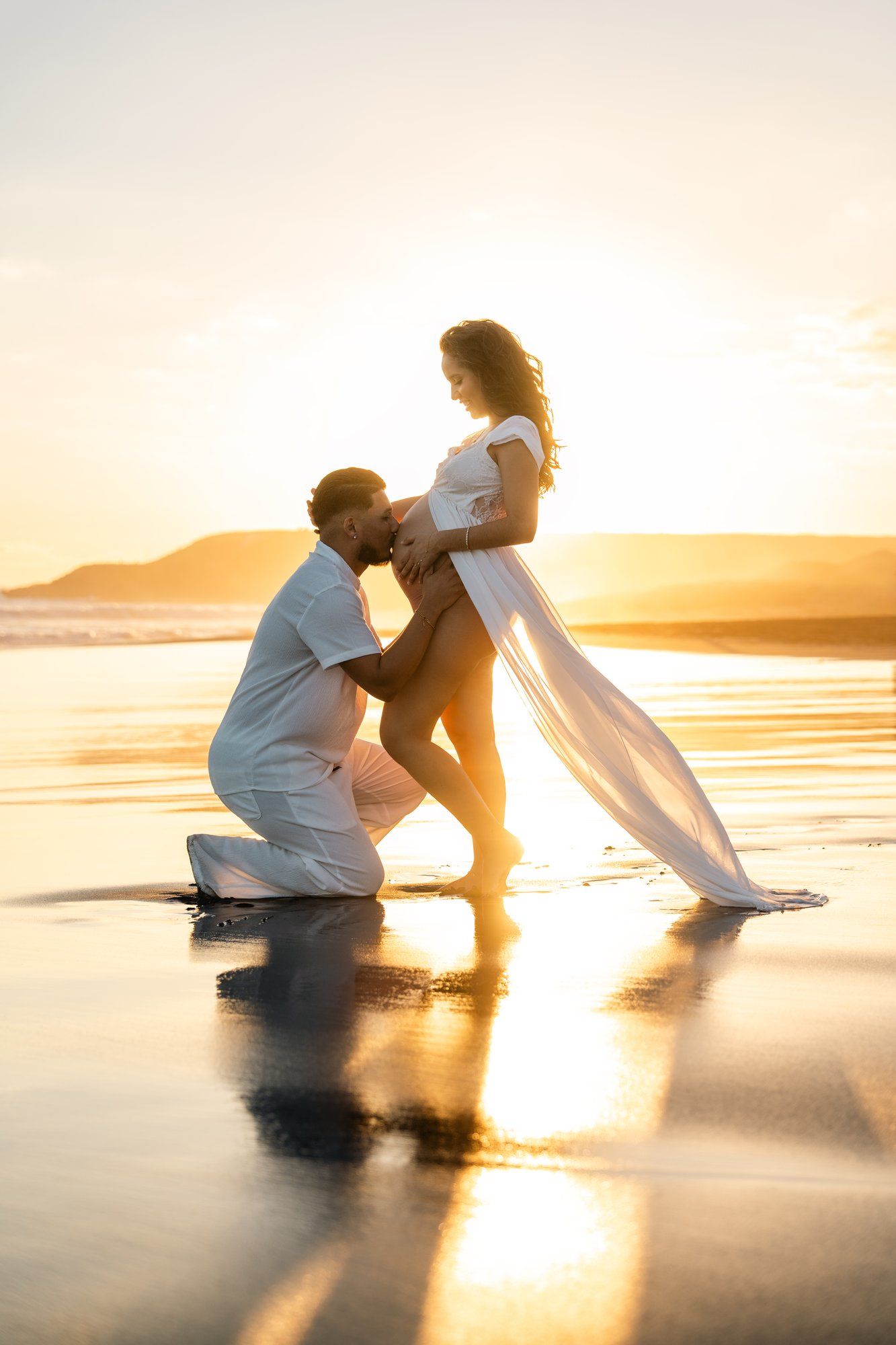 A couple in white clothing shares a tender moment on a reflective beach at golden sunset, with the man kneeling before a pregnant woman.
