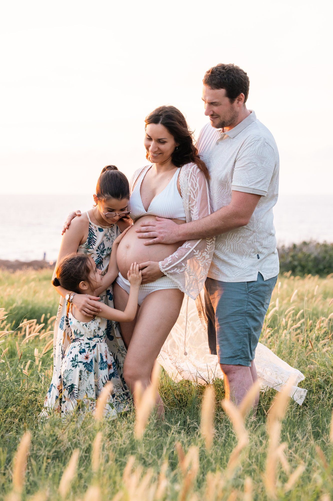 Pregnant woman in white lingerie and lace robe poses with family in golden field near ocean at sunset.