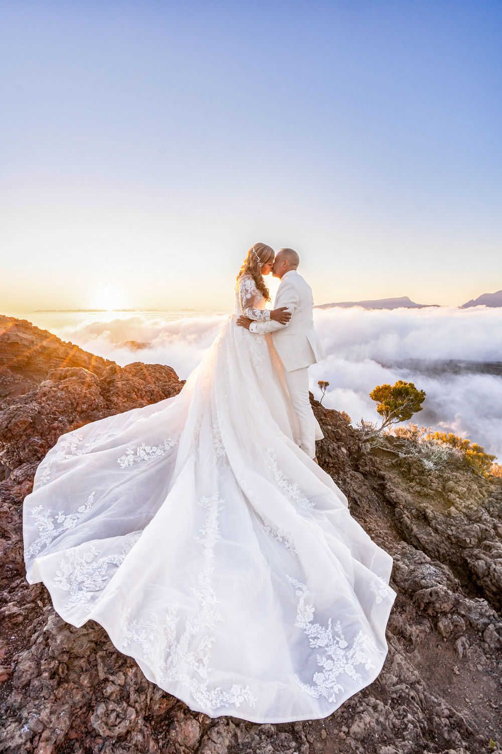 Un couple de mariés partage un moment intime au sommet d'une montagne au-dessus des nuages ​​au coucher du soleil, avec une robe blanche fl